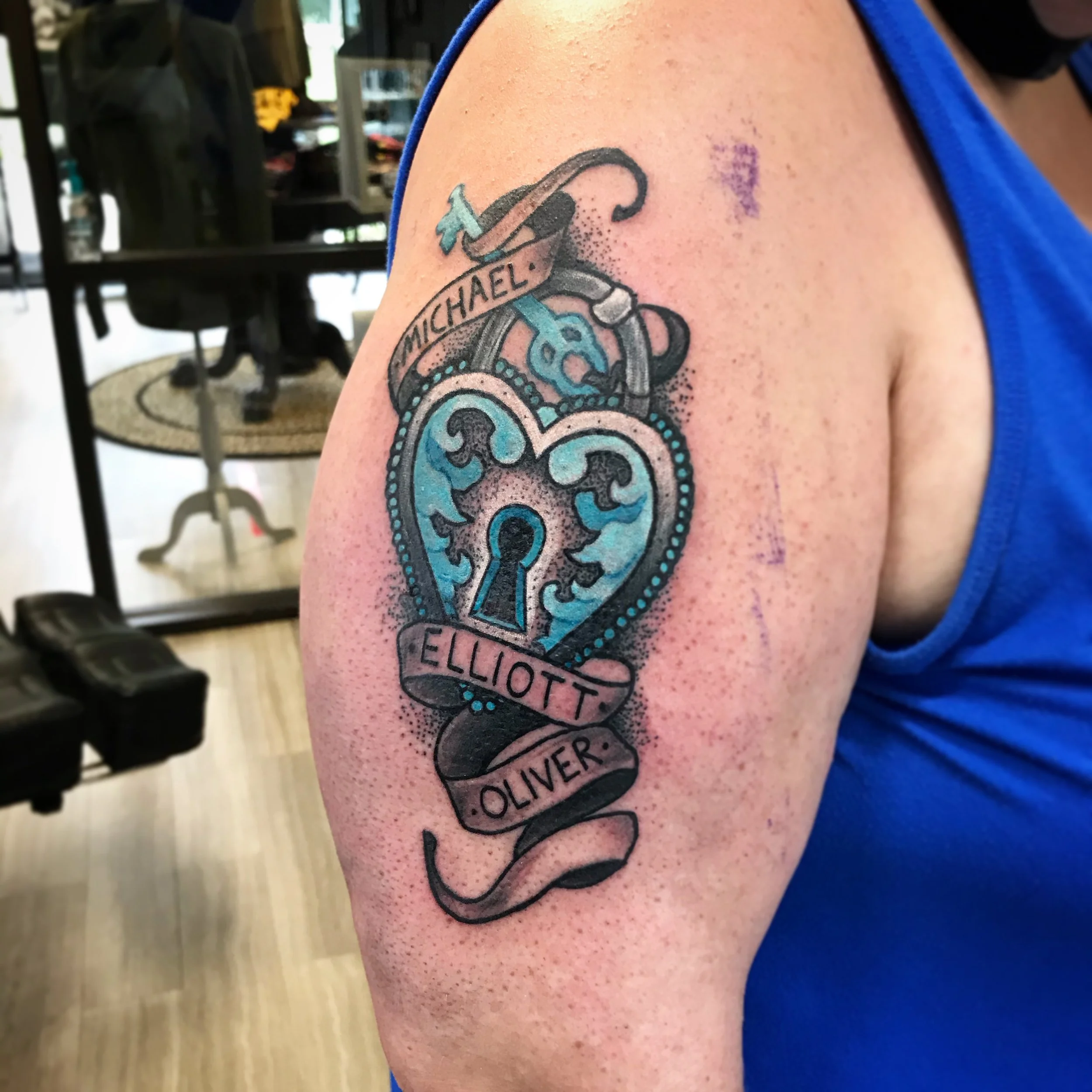 Tattoo of a heart-shaped lock with a keyhole and a banner with the names 'Michael,' 'Elliott,' and 'Oliver' on the person's upper arm.