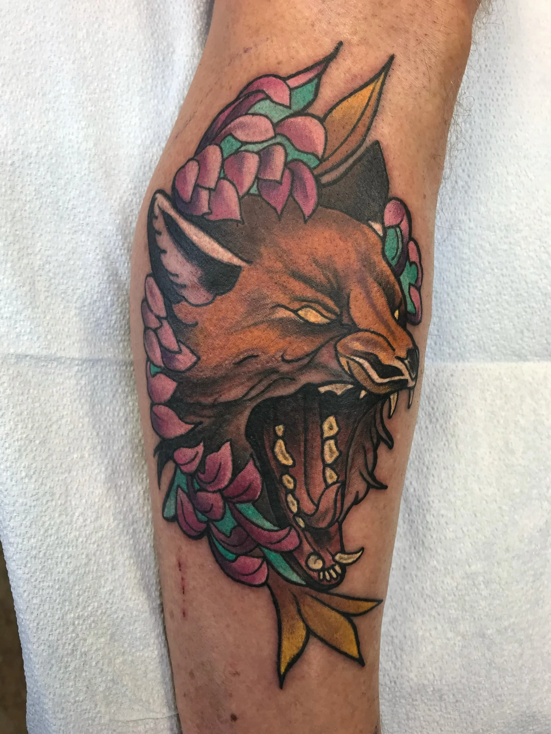 Tattoo of a roaring lion's head with open mouth, surrounded by pink flowers and green leaves on a person's arm.