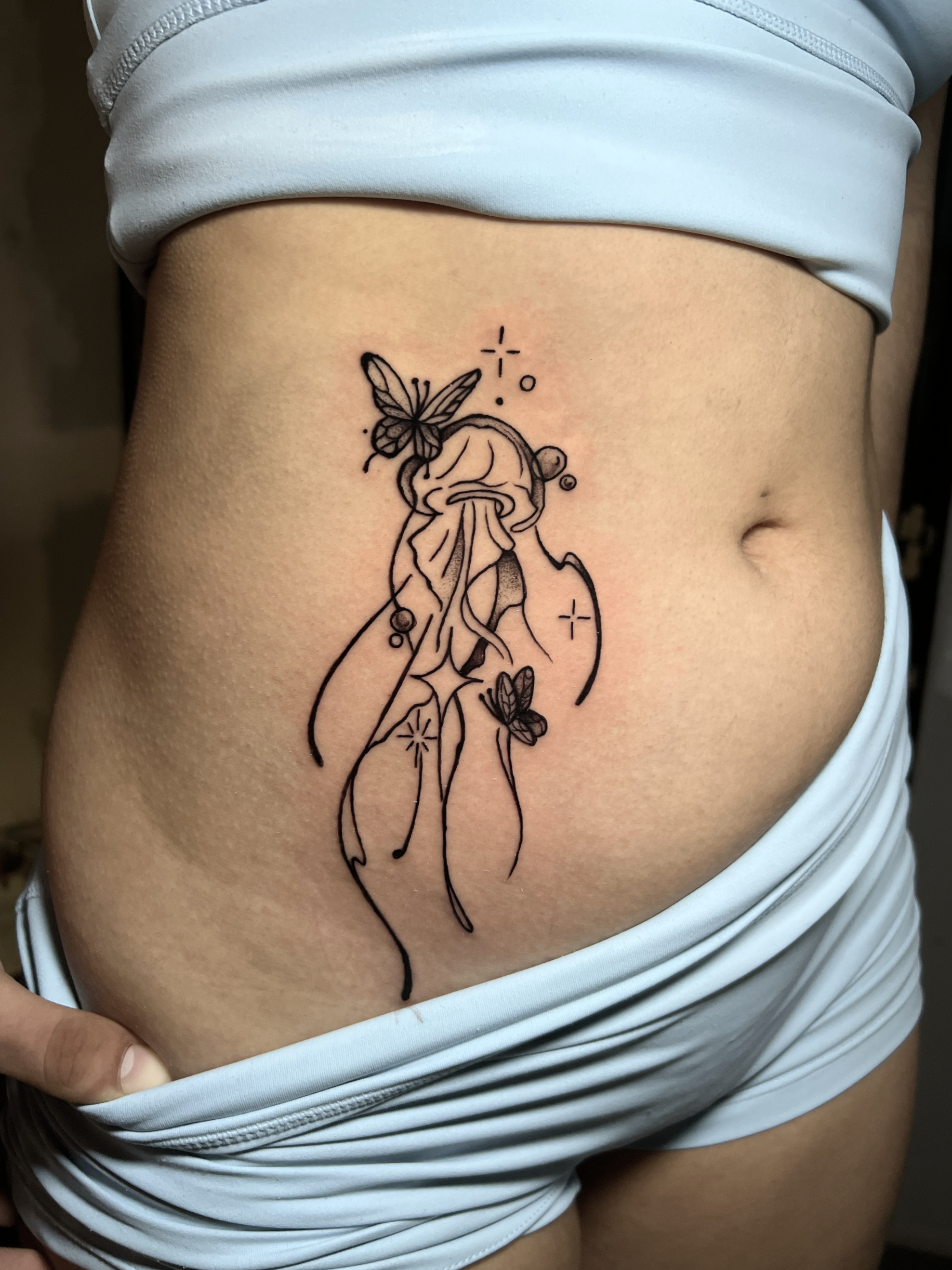 A tattoo on a person's side depicting a jellyfish with a butterfly above it and smaller butterflies around, with sparkles and dots for decorative effect.