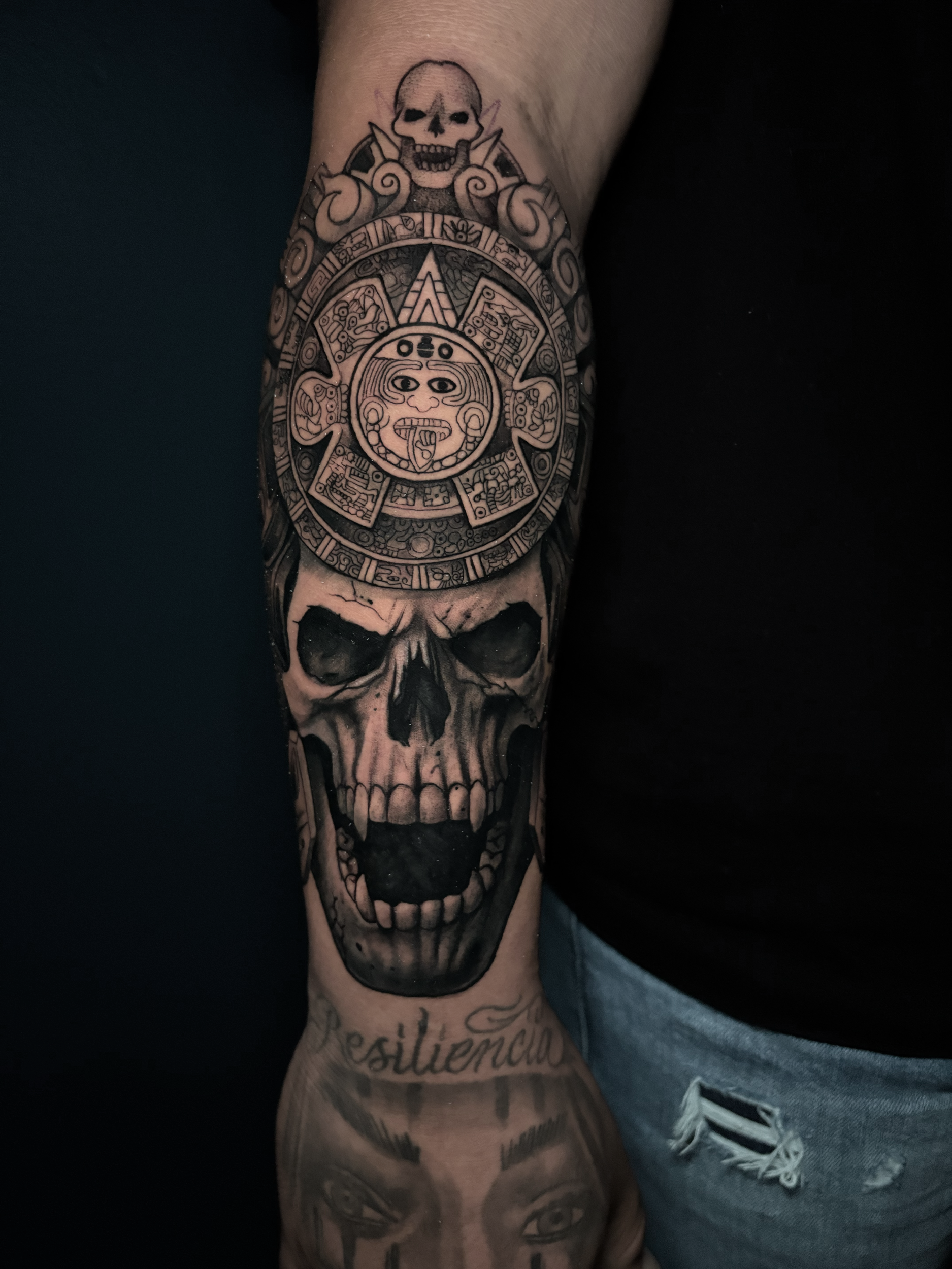 Black and gray tattoo sleeve featuring a skull at the top, Aztec calendar or sun motif in the middle, with a human face and a skull at the bottom, and text in cursive below.