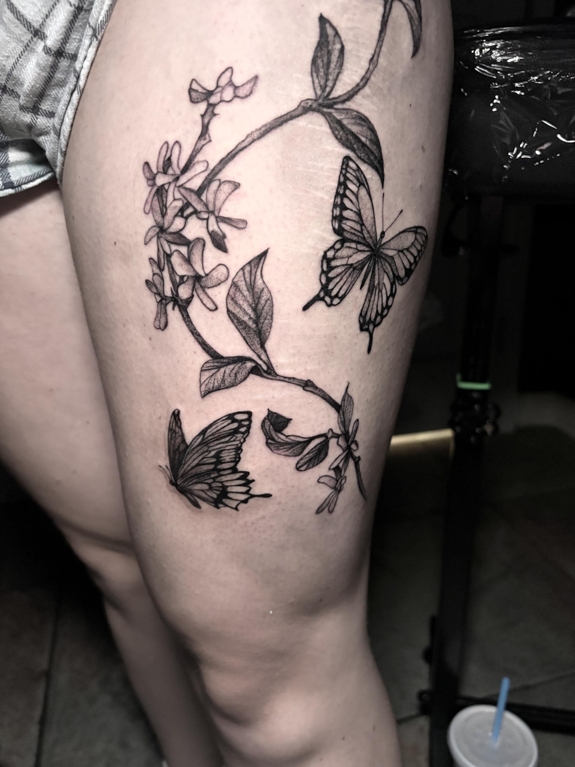 A person's thigh with a black ink tattoo of a butterfly and a branch with leaves and small flowers.