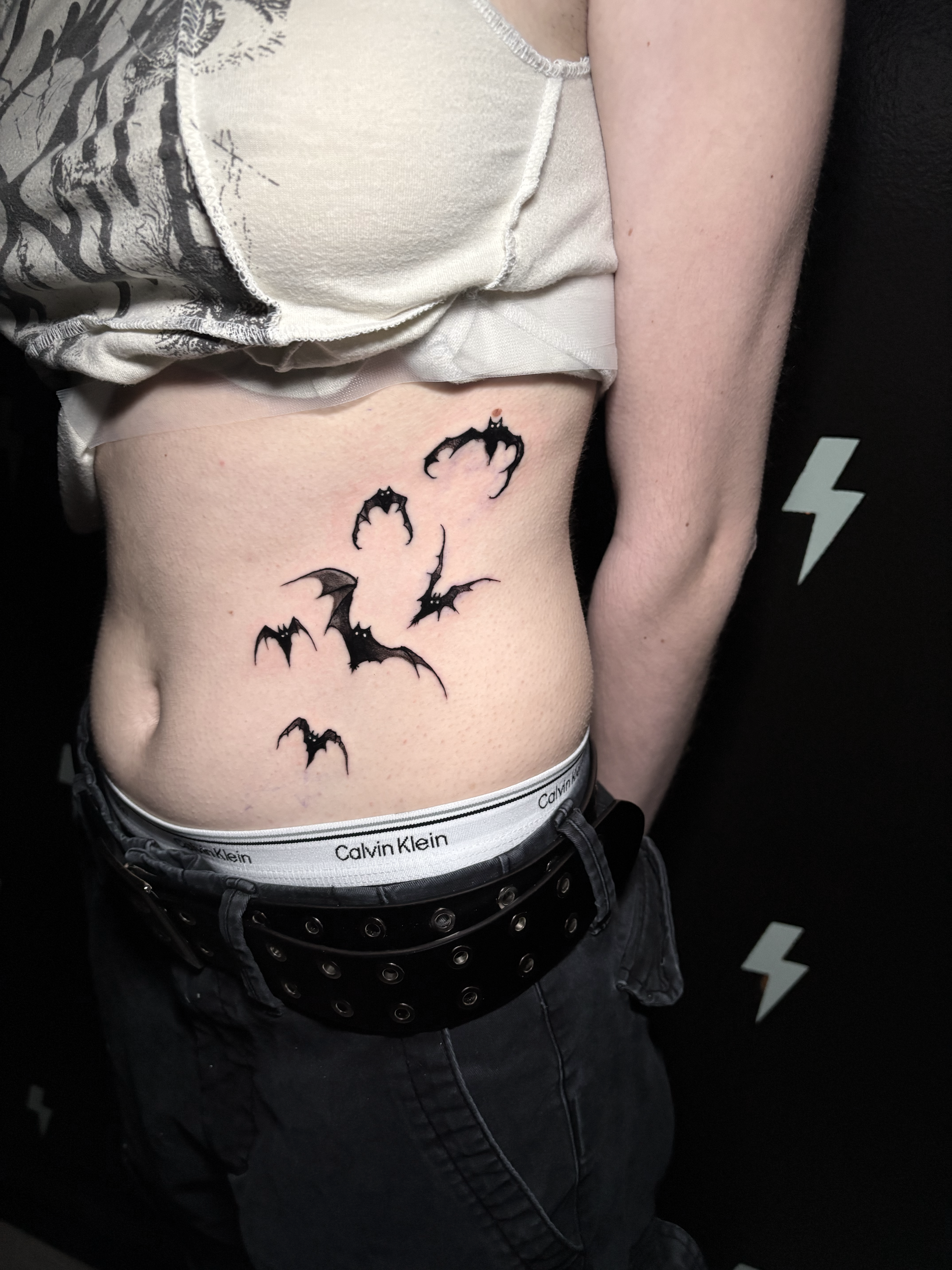 Close-up of a person's torso with a tattoo of black bats on the side of their ribcage, wearing black pants with a studded belt and Calvin Klein underwear waistband visible, against a black background with white lightning bolt graphics.