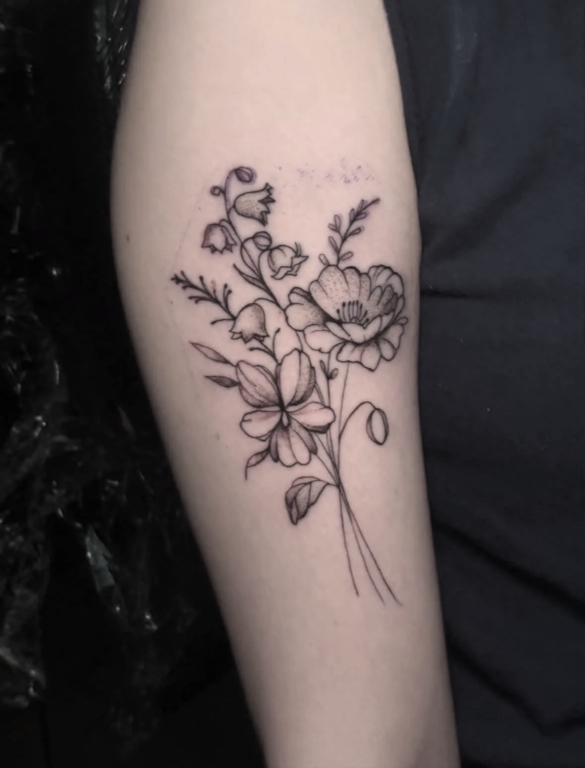Black outline floral tattoo on person's arm, featuring various flowers with detailed petals and leaves.