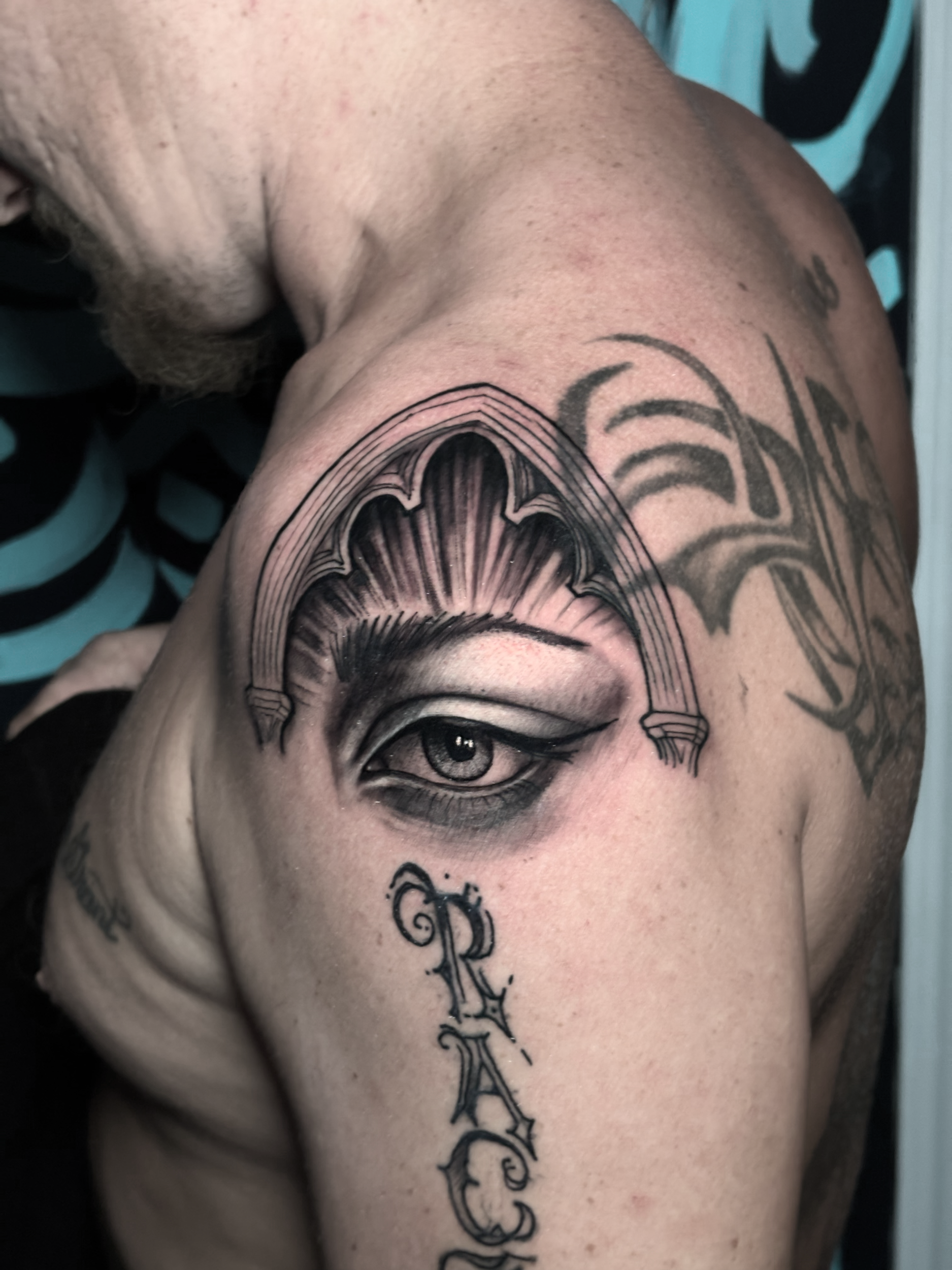 Close-up of a person's shoulder with a detailed black and gray tattoo of an eye and part of a face, featuring intricate shading and line work.