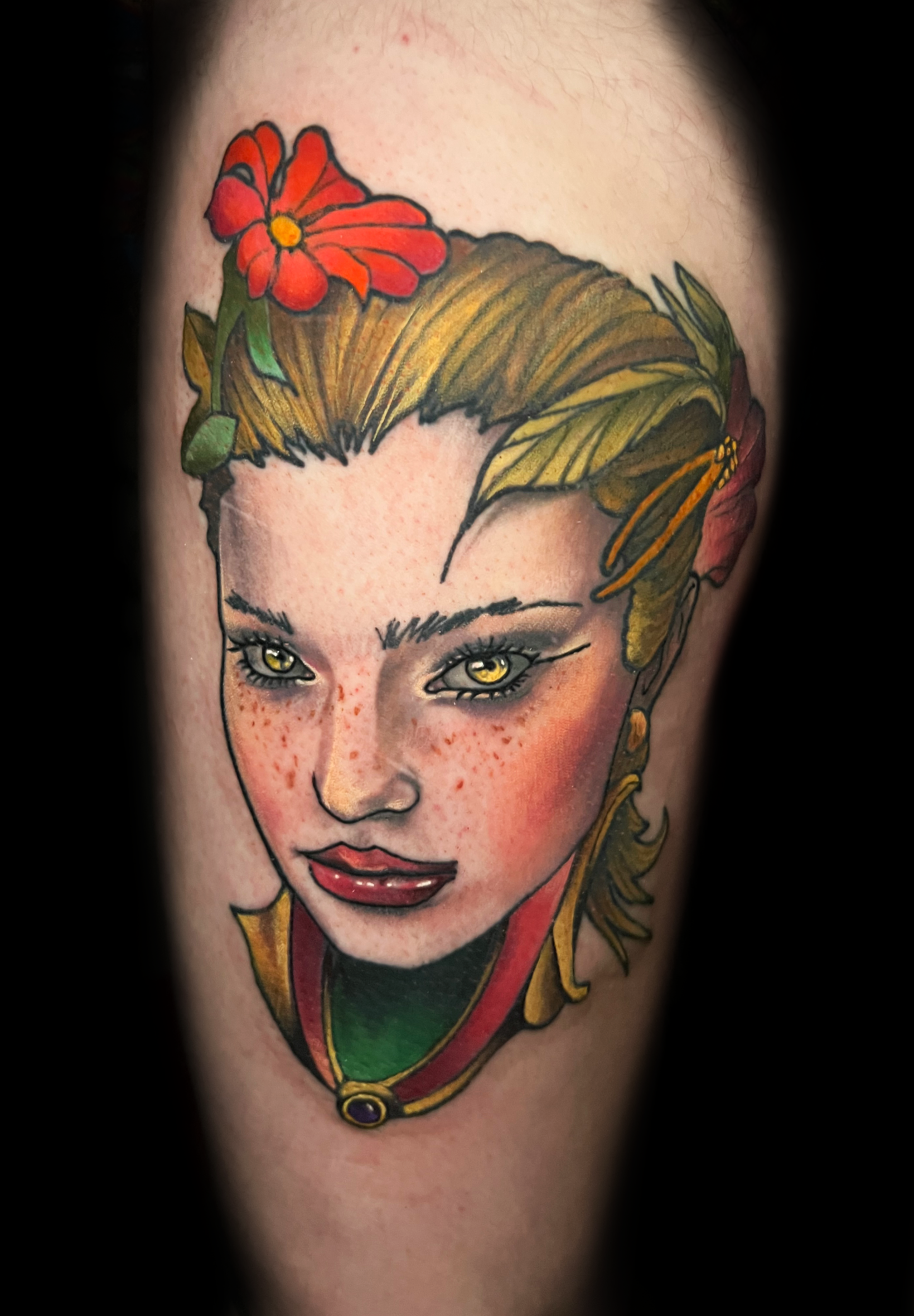 Close-up of a colorful tattoo of a woman's face with blonde hair, yellow eyes, freckles, and a red flower with green leaves and a gold necklace on the forehead.