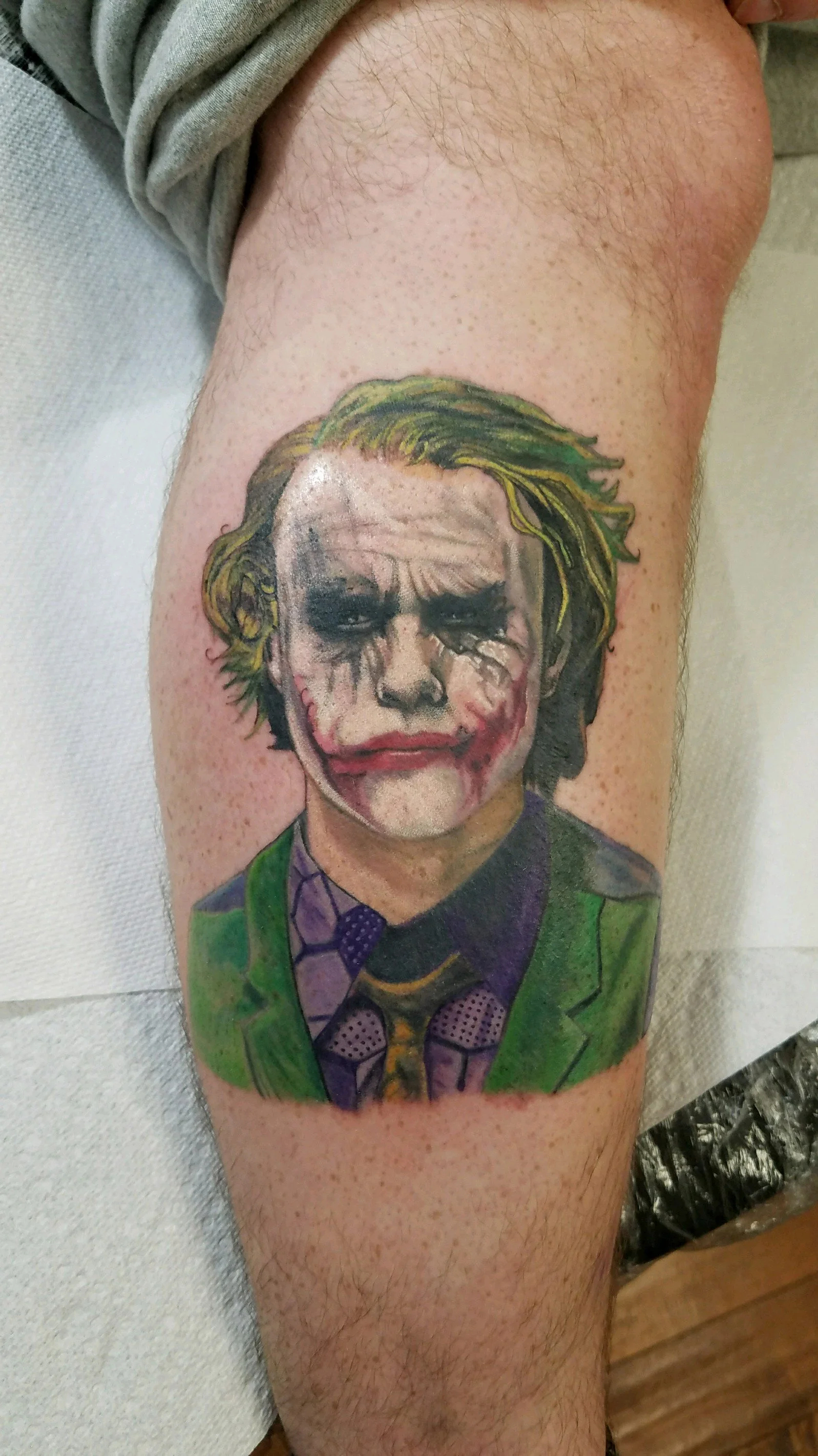 Tattoo of the Joker from Batman on a person's leg, showing his face with messy green hair, white face paint, black eye makeup, and a red-painted smile, wearing a green suit, purple shirt, and patterned tie.