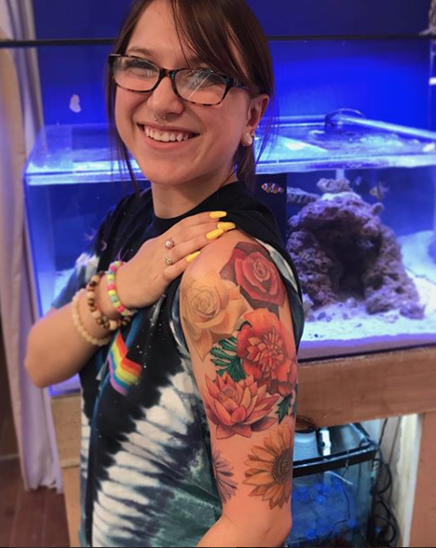 woman with glasses and rainbow-painted nails showing colorful flower tattoo on her upper arm in front of an aquarium