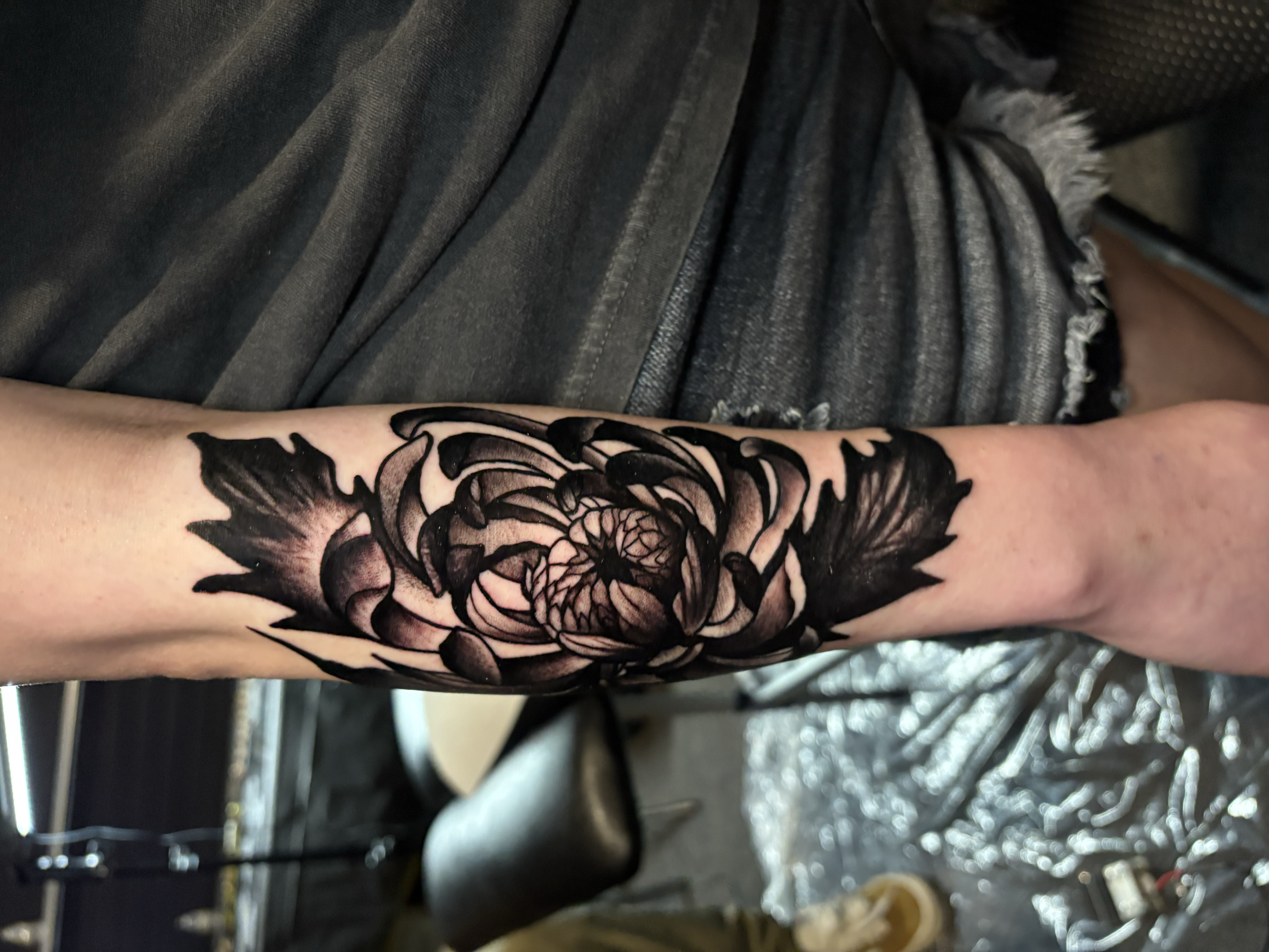 Close-up of a person's arm and hand with a large floral black tattoo, wearing a black and gray striped shirt, in a store or workshop setting.