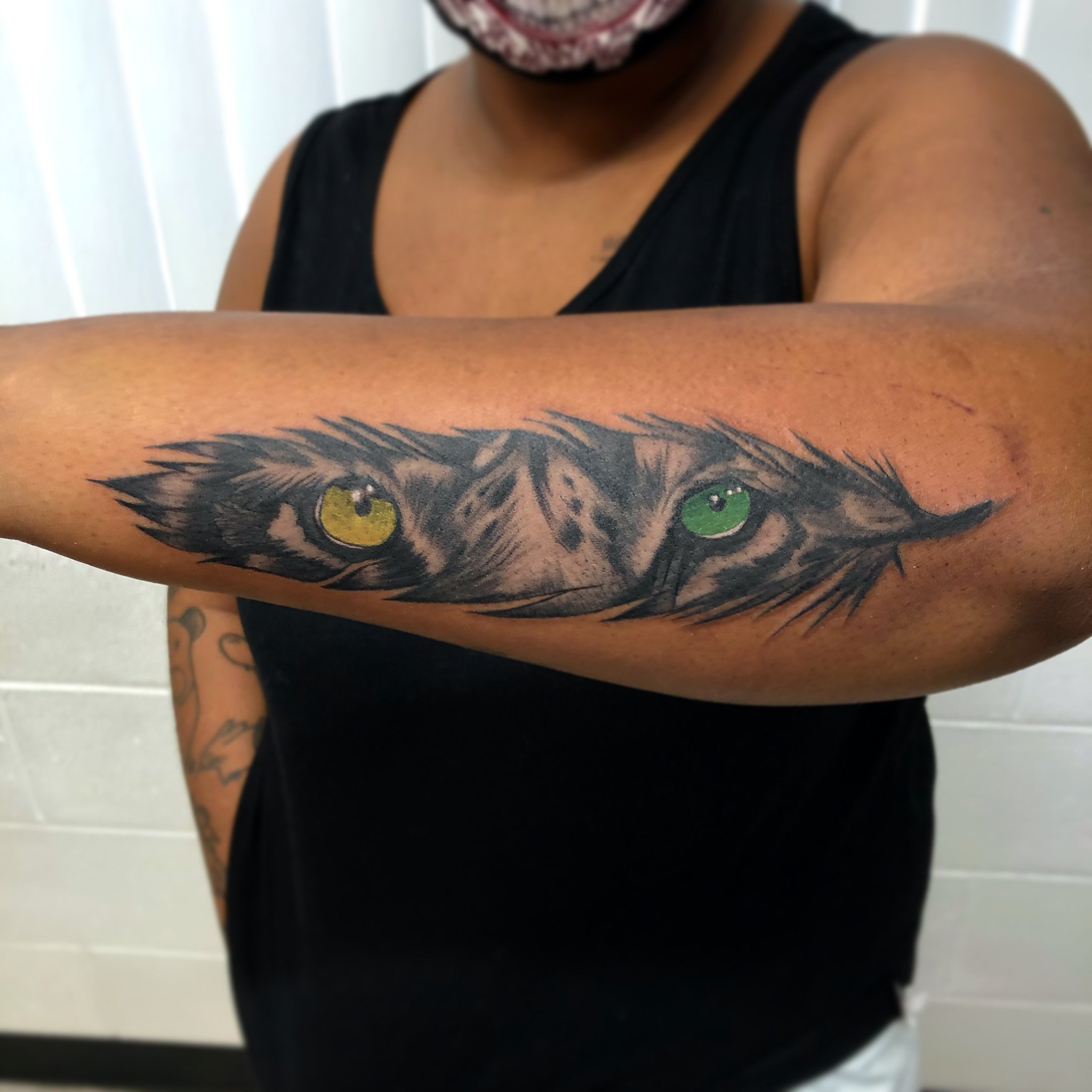 Tattoo of a feline's face with yellow and green eyes on a person's forearm.