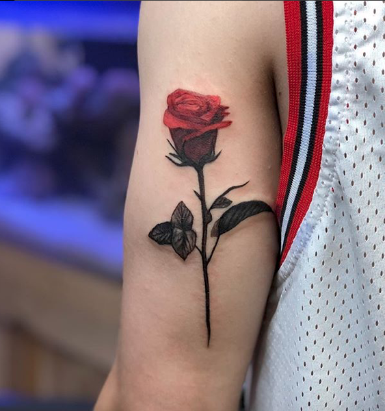 Tattoo of a red rose with green leaves on a person's upper arm near the shoulder, wearing a sleeveless sports jersey.
