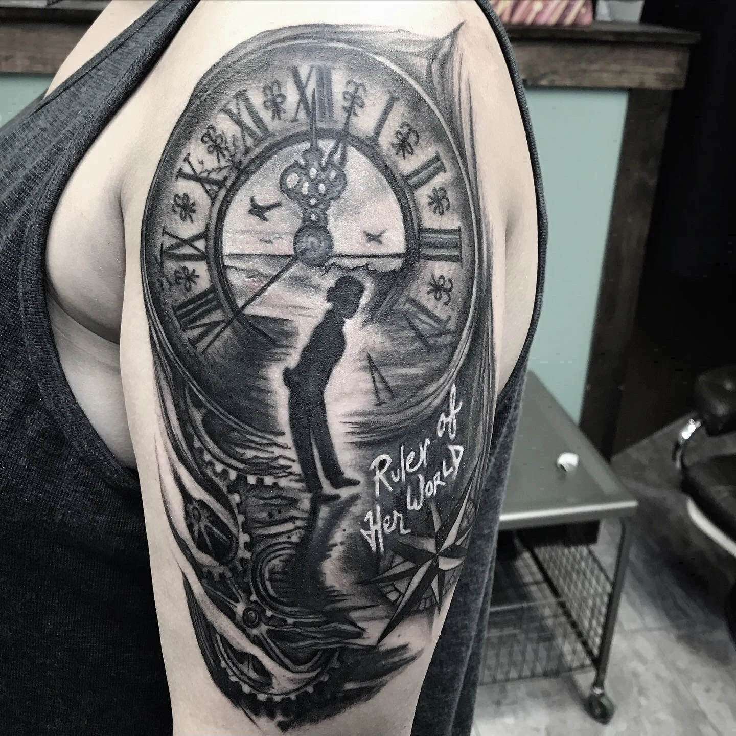 A tattoo of a clock with Roman numerals, a woman's silhouette, ocean scene, gears, and a compass on a person's upper arm. The tattoo includes the phrase "Roller of Her War".
