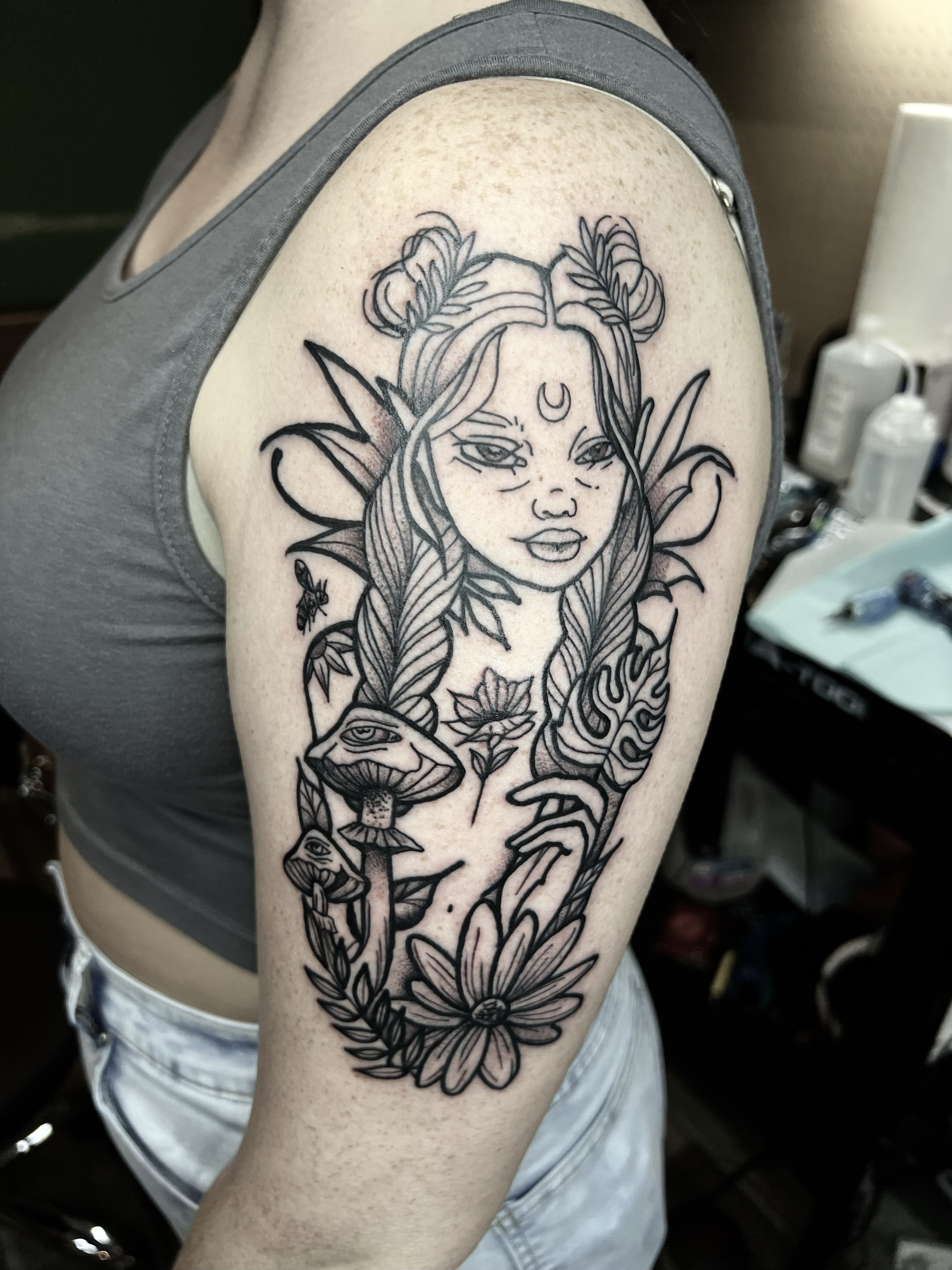 Black fine line micro outline tattoo of a fantasy-style female elf with pointed ears, long wavy hair styled in two buns, a crescent moon on her forehead, and surrounded by mushrooms, a flower, a bee, and decorative foliage, on a person's upper arm.