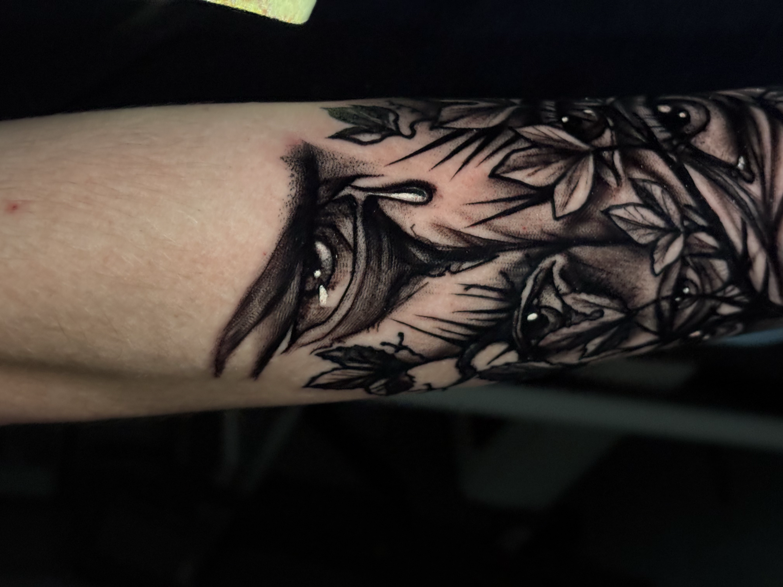 Close-up photo of a person's tattooed arm, featuring black ink floral and leaf designs on light skin.