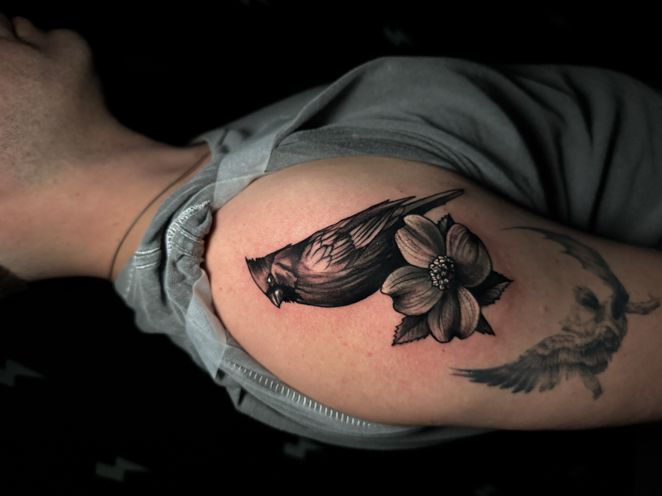Close-up of a person's torso, showing a detailed black and gray floral tattoo on the upper arm, with gray clothing partially visible.
