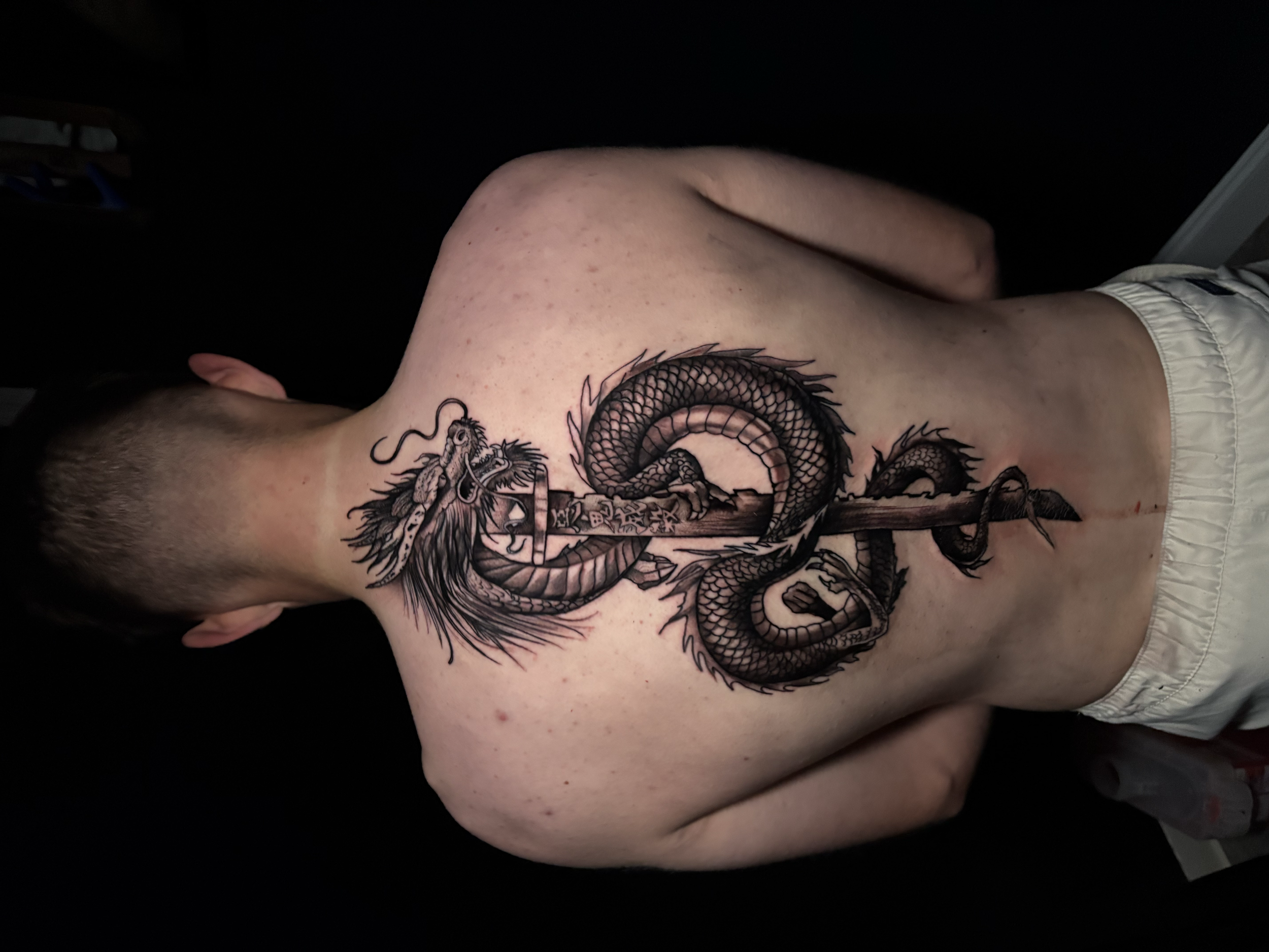 A person with a detailed black and gray dragon tattoo on their upper arm and shoulder, wearing light-colored shorts, with a dark background.