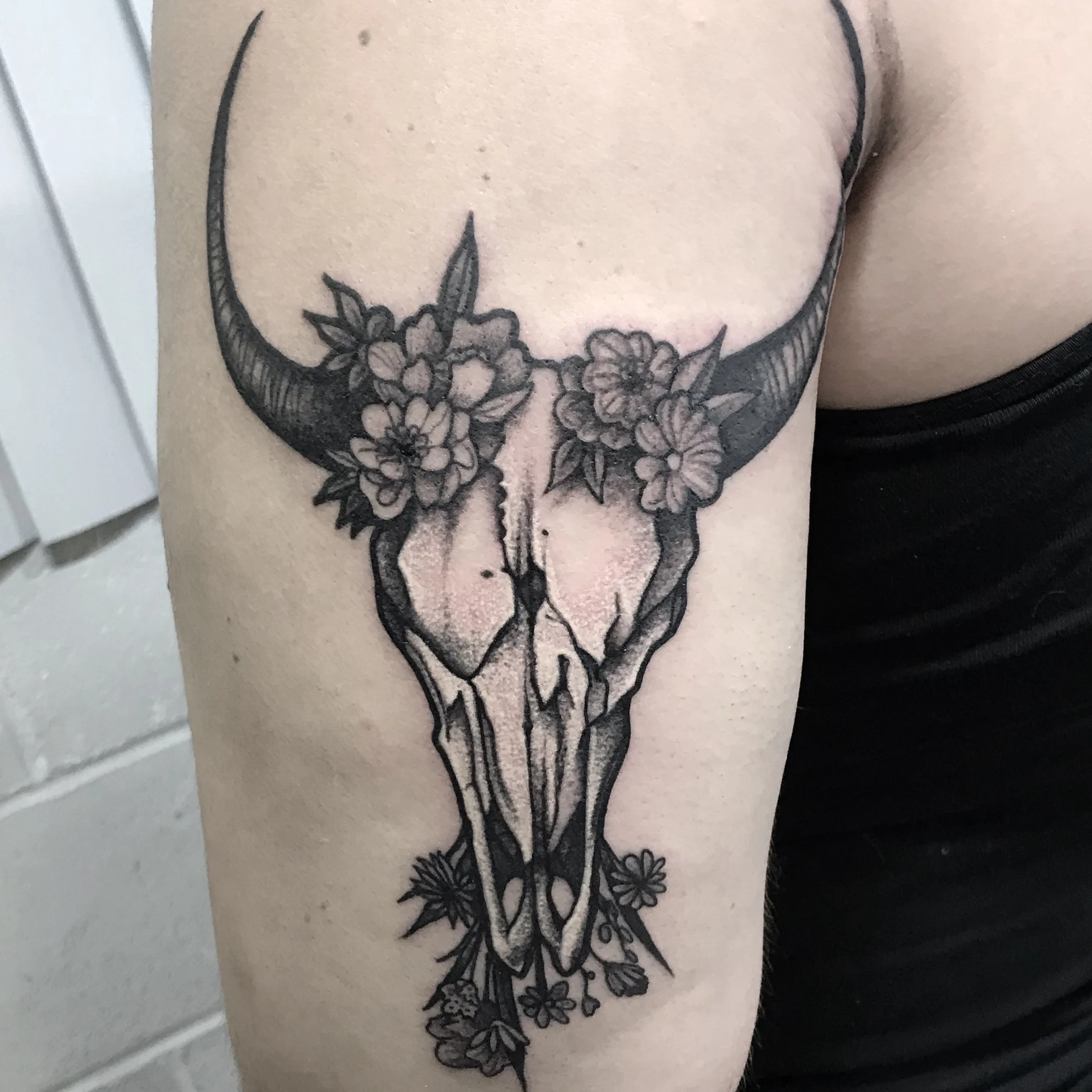 Black and gray tattoo of a deer skull with flowers and leaves around it on a person's upper arm.