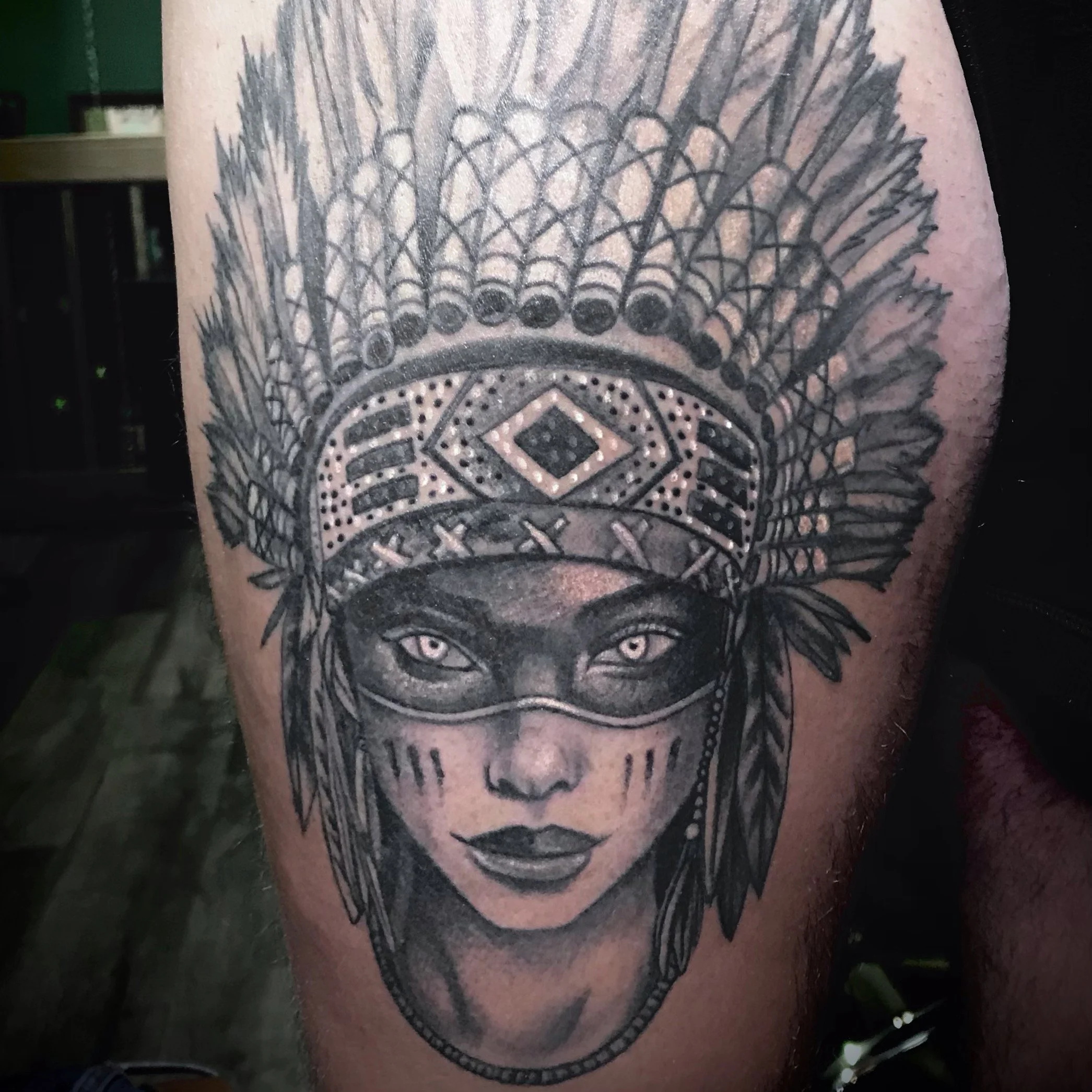 Tattoo of a woman with dark skin, wearing a Native American headdress with detailed feathers, and a face mask covering her eyes.