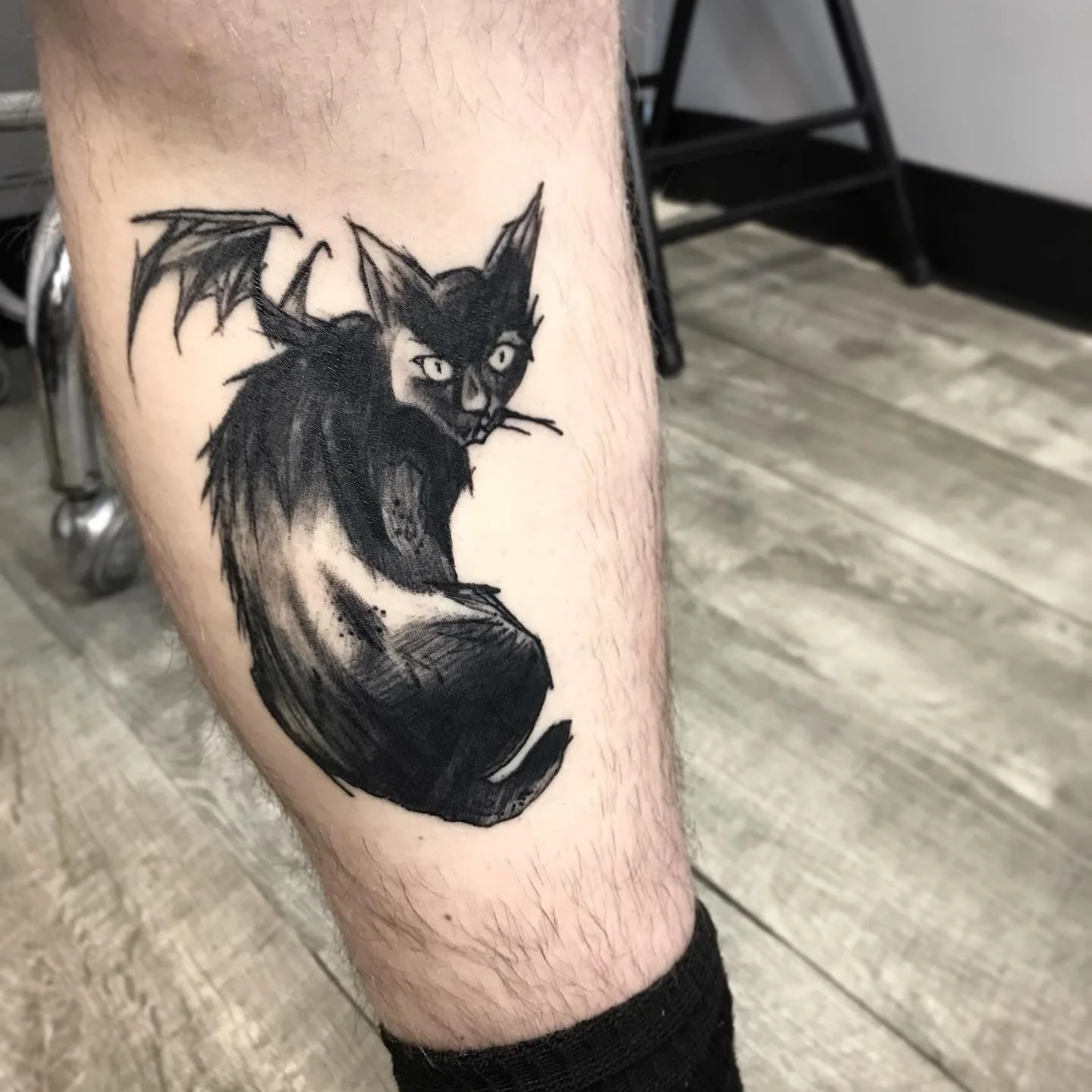 Black and gray tattoo of a mythical creature with cat-like face, bat wings, and a long tail on person's leg