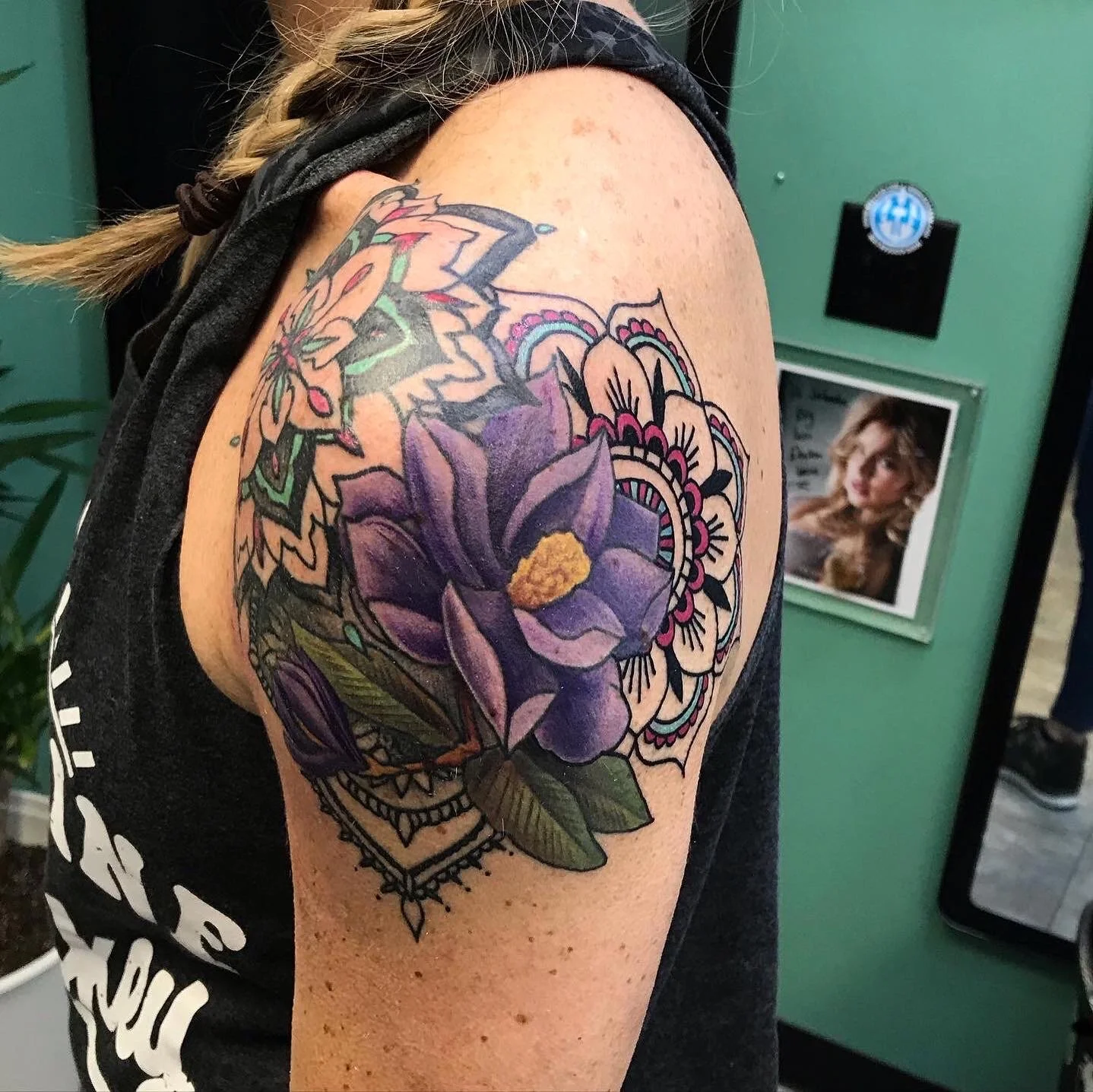 Tattoo of a large purple flower with yellow center on a person's upper arm, surrounded by intricate black and pink mandala-like designs and green leaves.