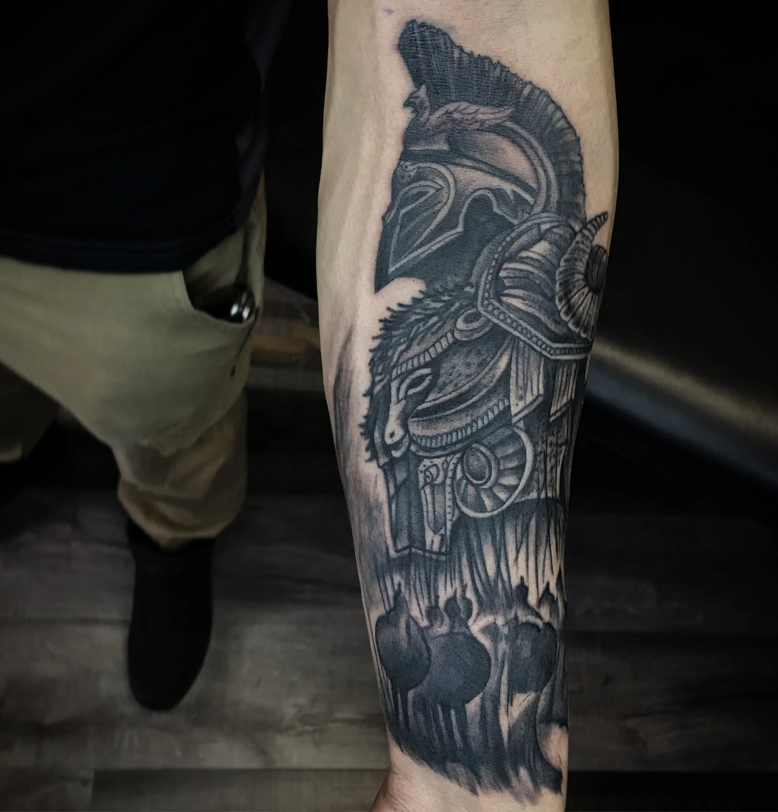 Black and gray tattoo of a Greek warrior helmet, horse's head, and torches on a person's arm.
