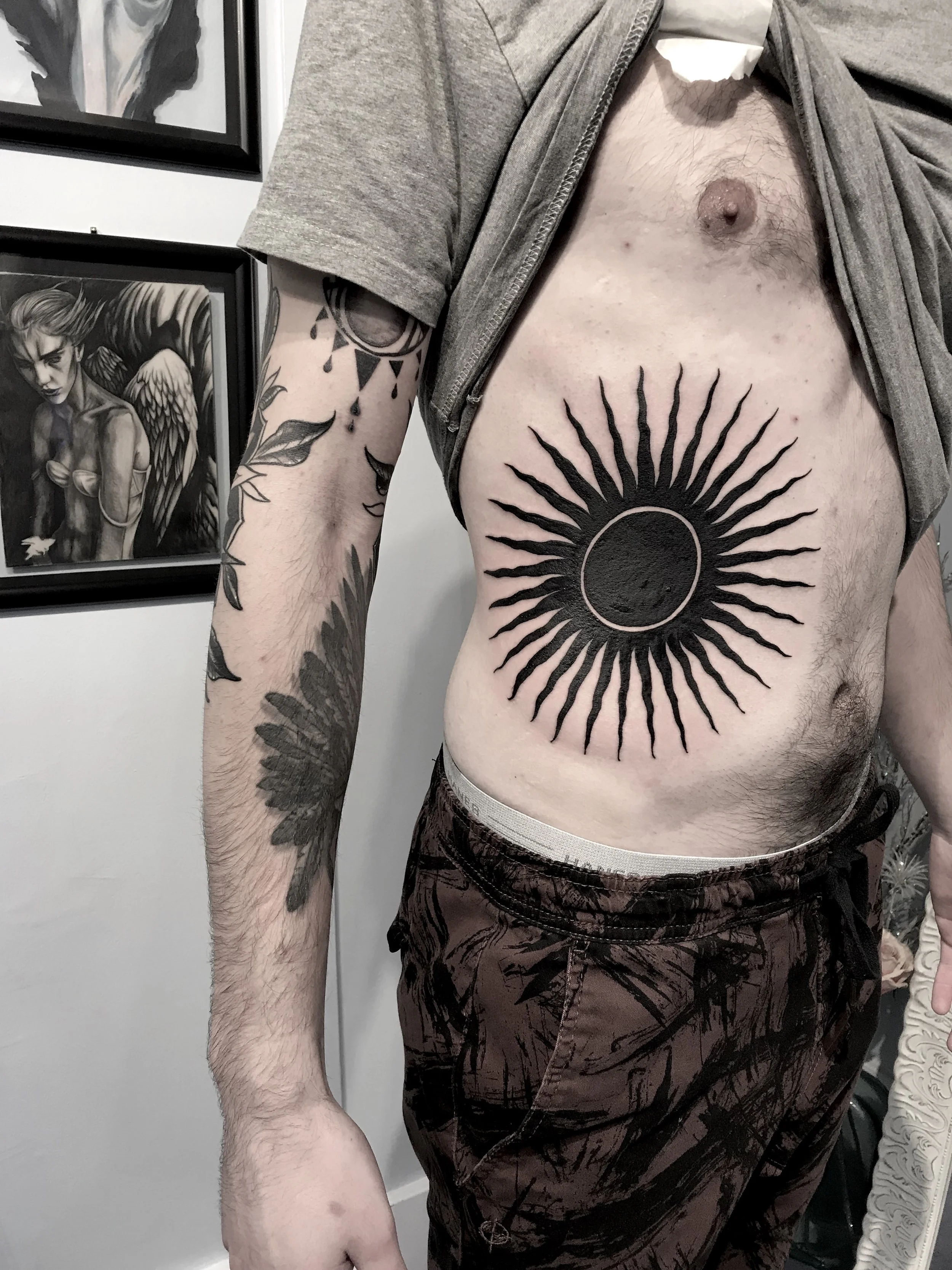 A man with tattoos on his arm and torso, lifting his shirt to reveal a large black tattoo of a sun with rays and a circle in the center on his chest.