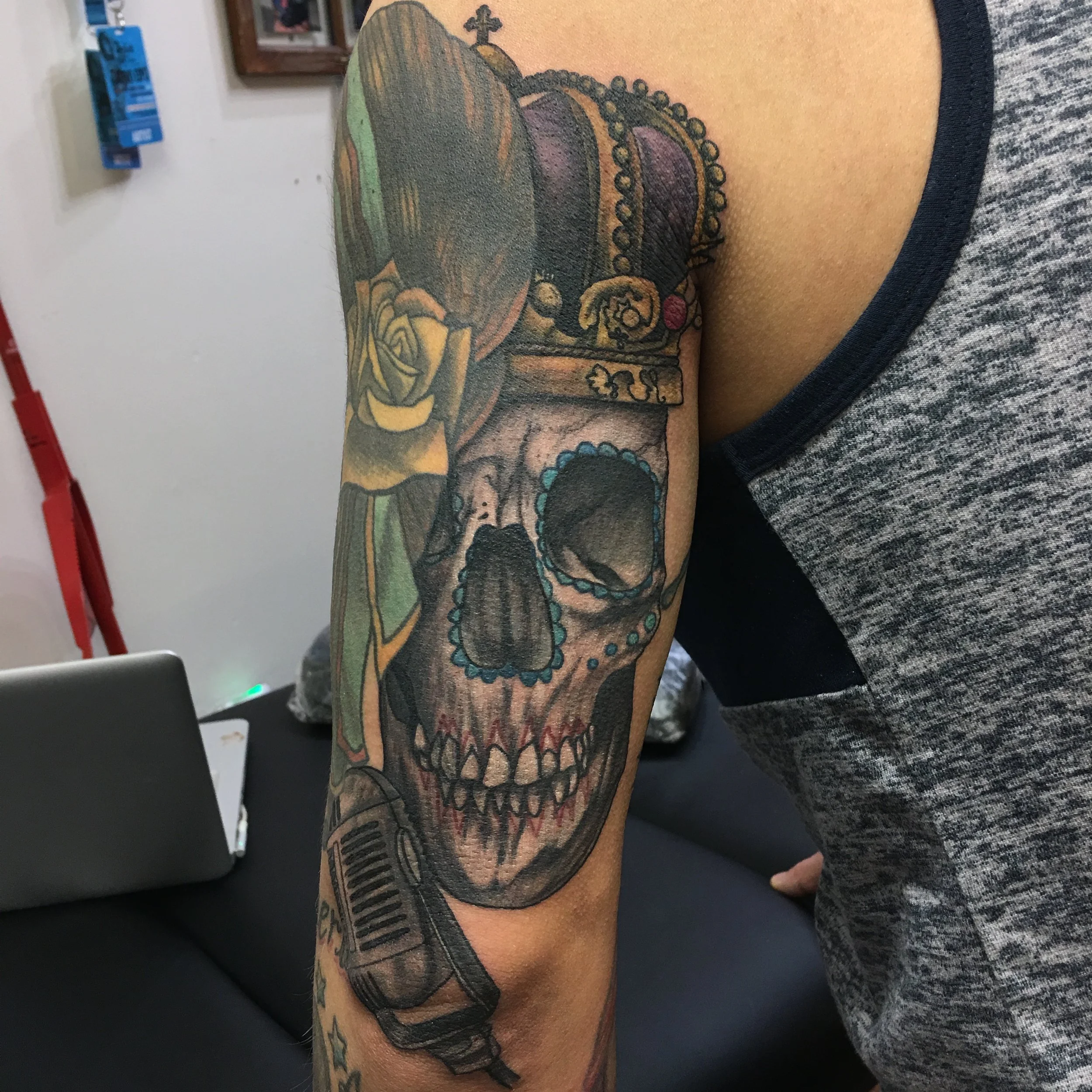 Tattoo of a skull wearing a crown, with a yellow rose and a gun on the arm, on a person's upper arm.