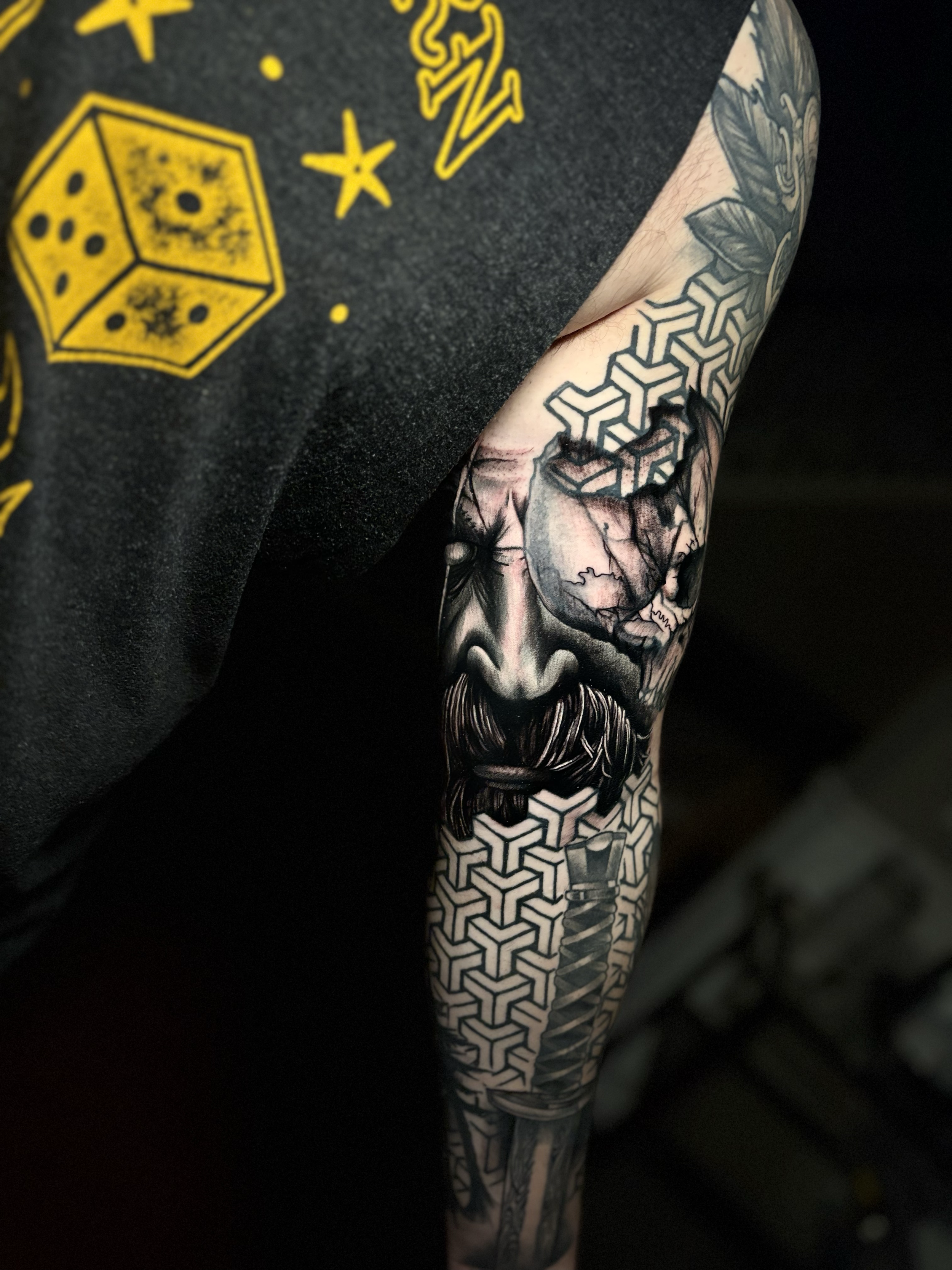 Close-up of a tattooed arm with geometric and floral designs, partially covered by a black shirt with yellow graphic prints.