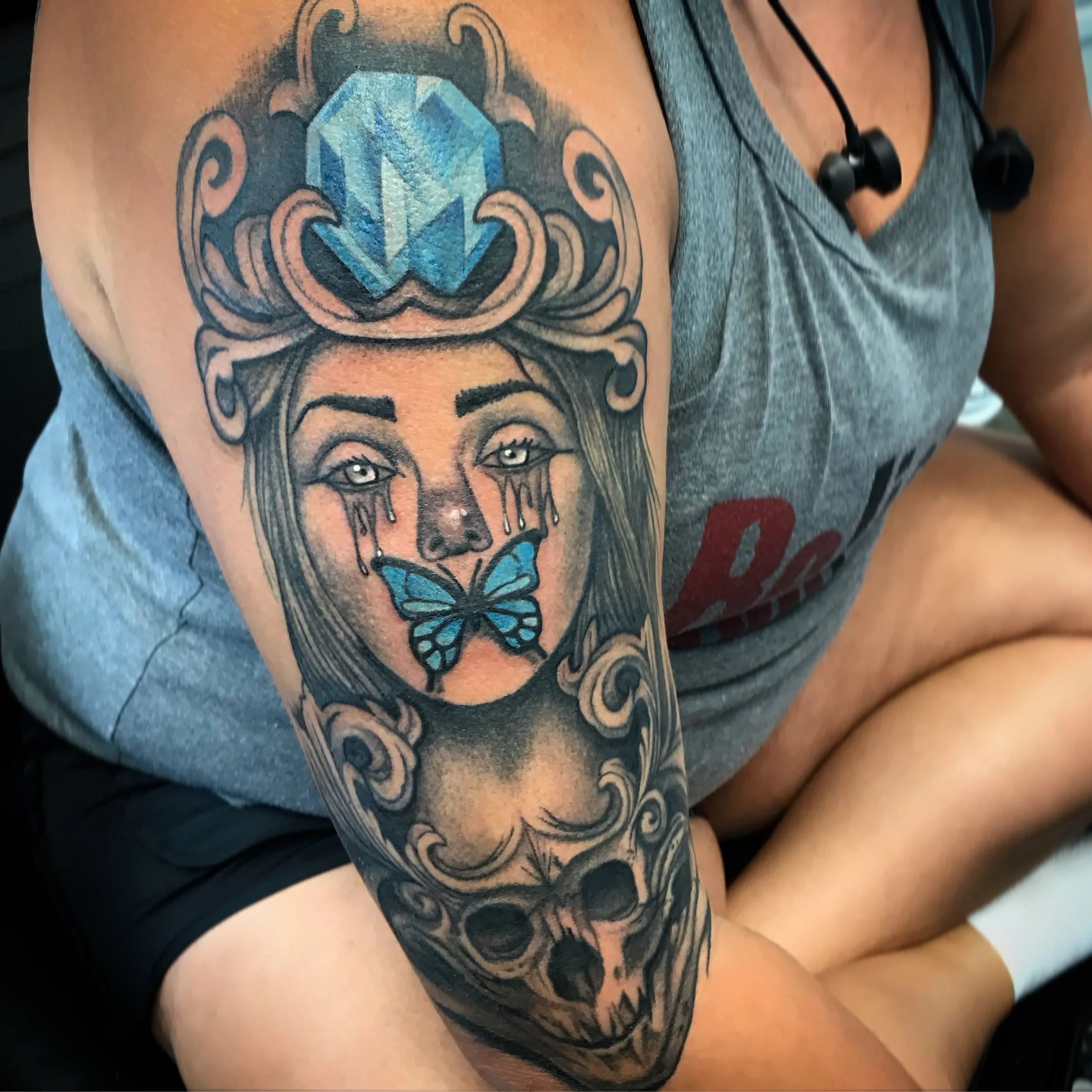Tattoo of a woman's face with tears, a butterfly near her lips, and a skull at the bottom on a person's arm.