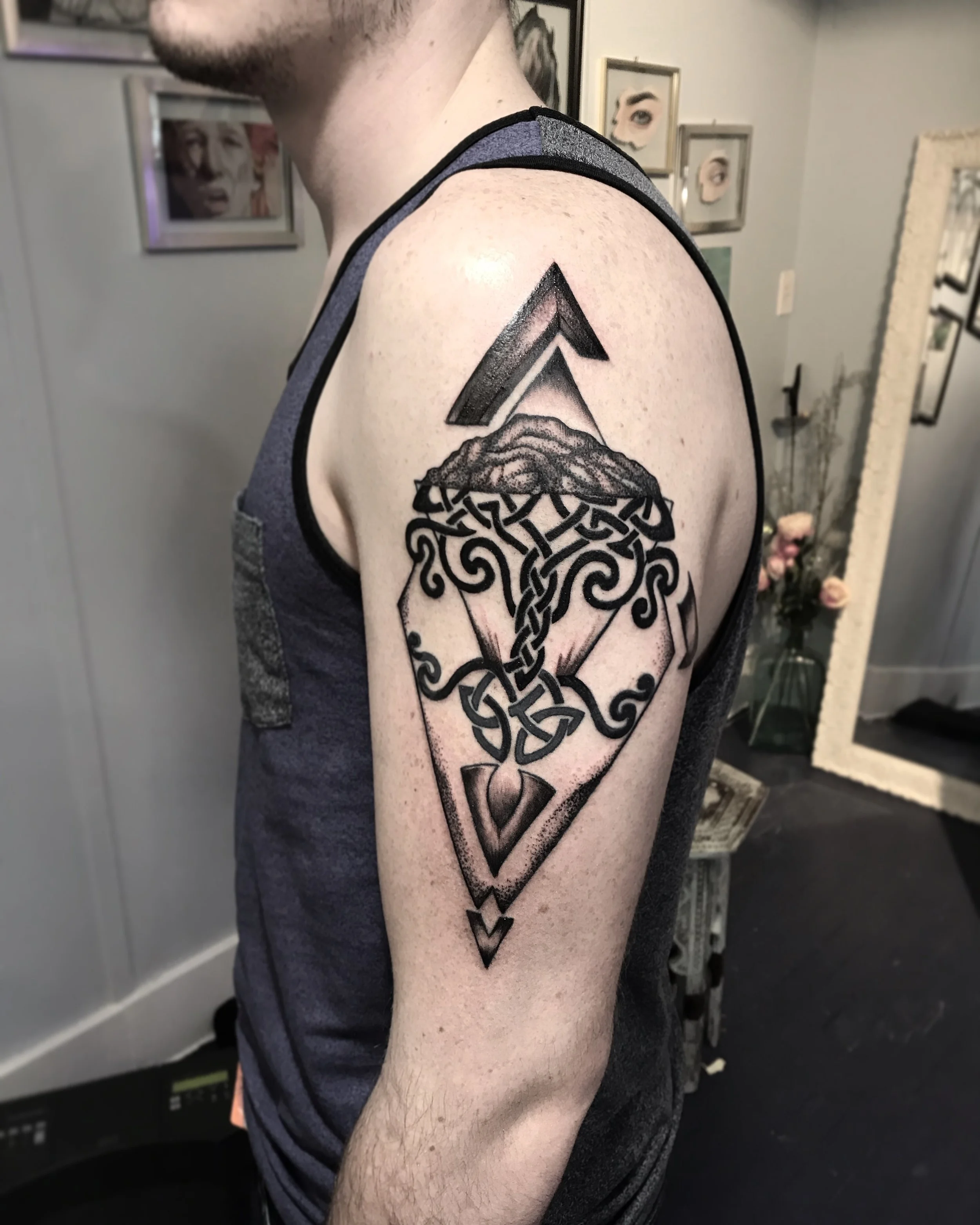 Black and gray geometric tattoo on a person's upper arm, featuring an arrow pointing upward, intertwined Celtic knots, and a diamond shape.