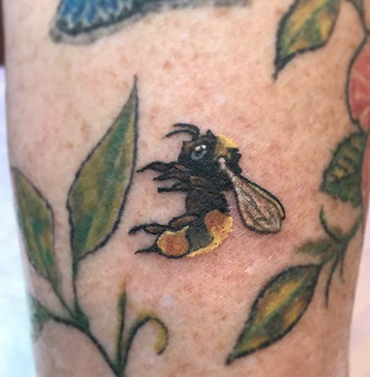 Tattoo of a bee surrounded by green leaves on skin.