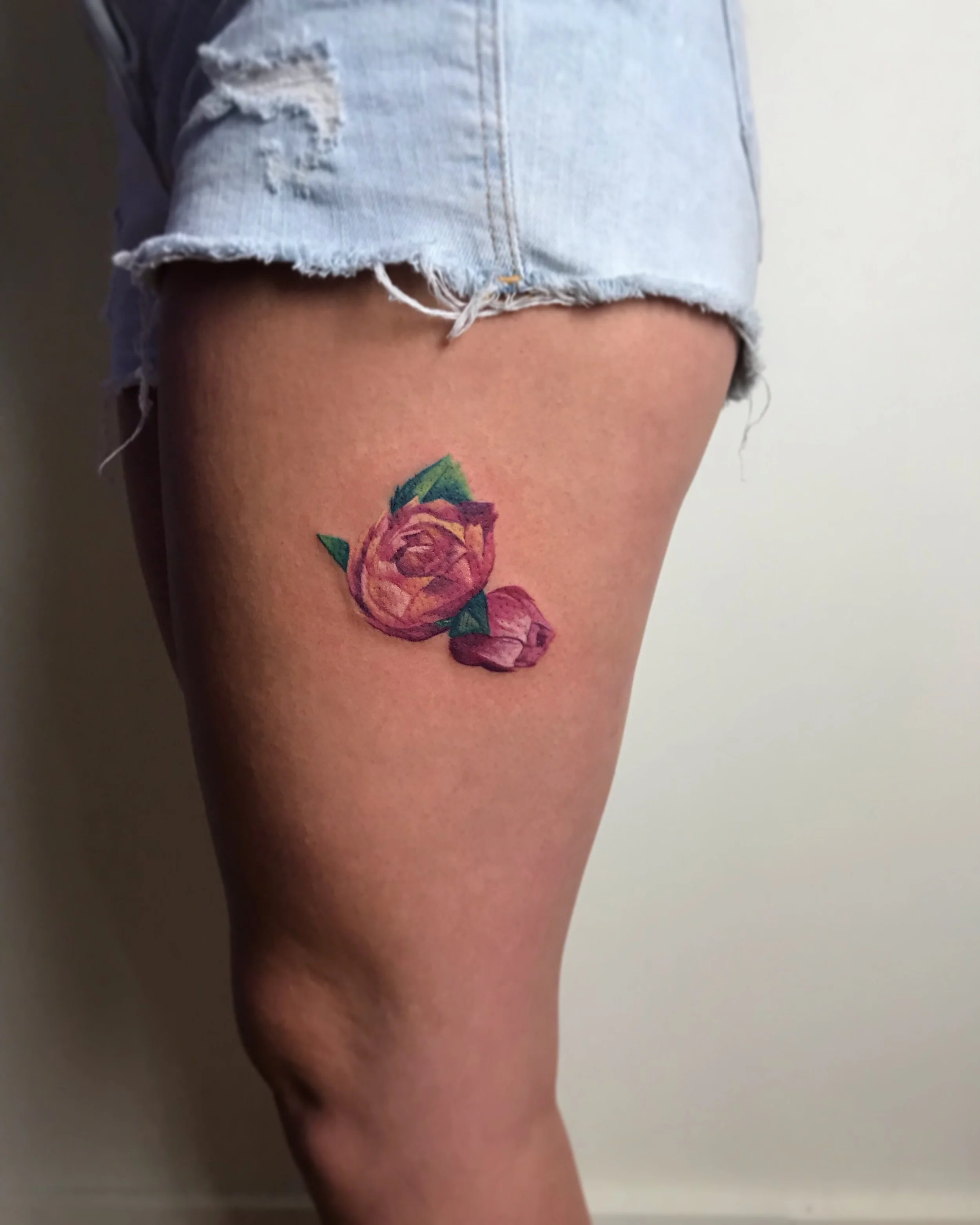 Tattoo of a pink and orange rose with green leaves on a person's upper thigh, partially covered by ripped denim shorts.
