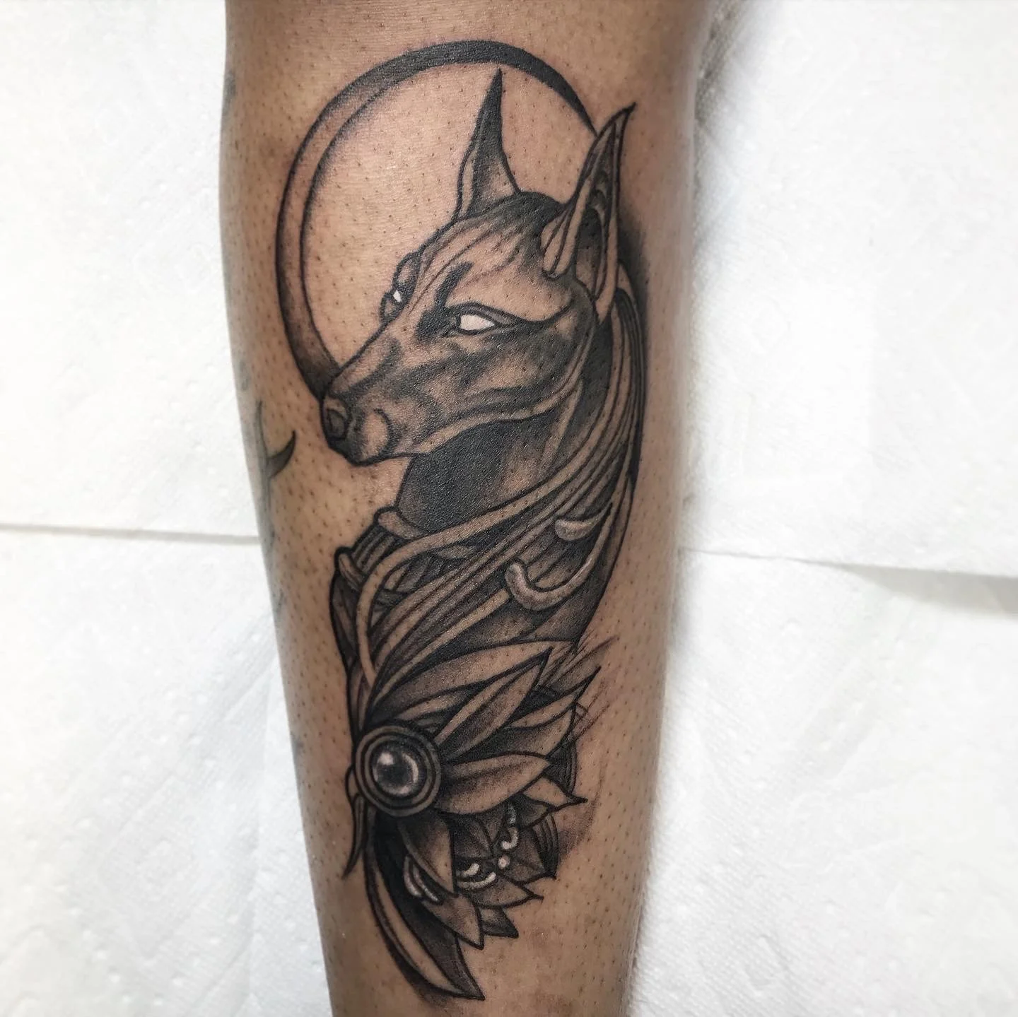 Black and gray tattoo of a German Shepherd dog with a moon behind its head, surrounded by ornamental designs.