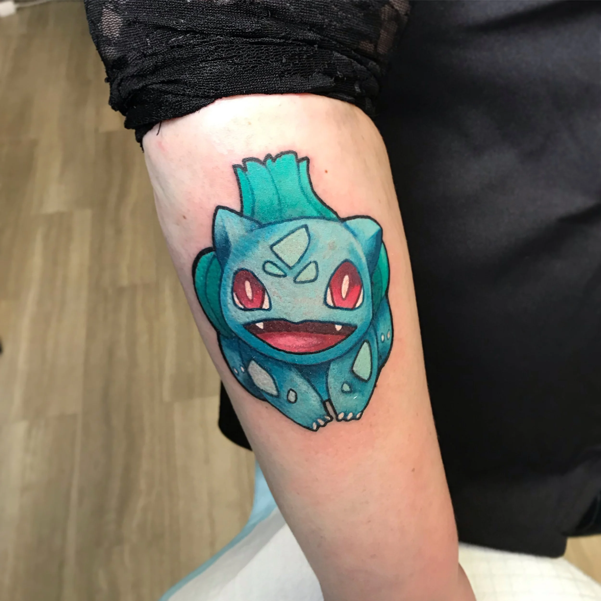 Colorful tattoo of Bulbasaur, a Pokémon, on person's upper arm.