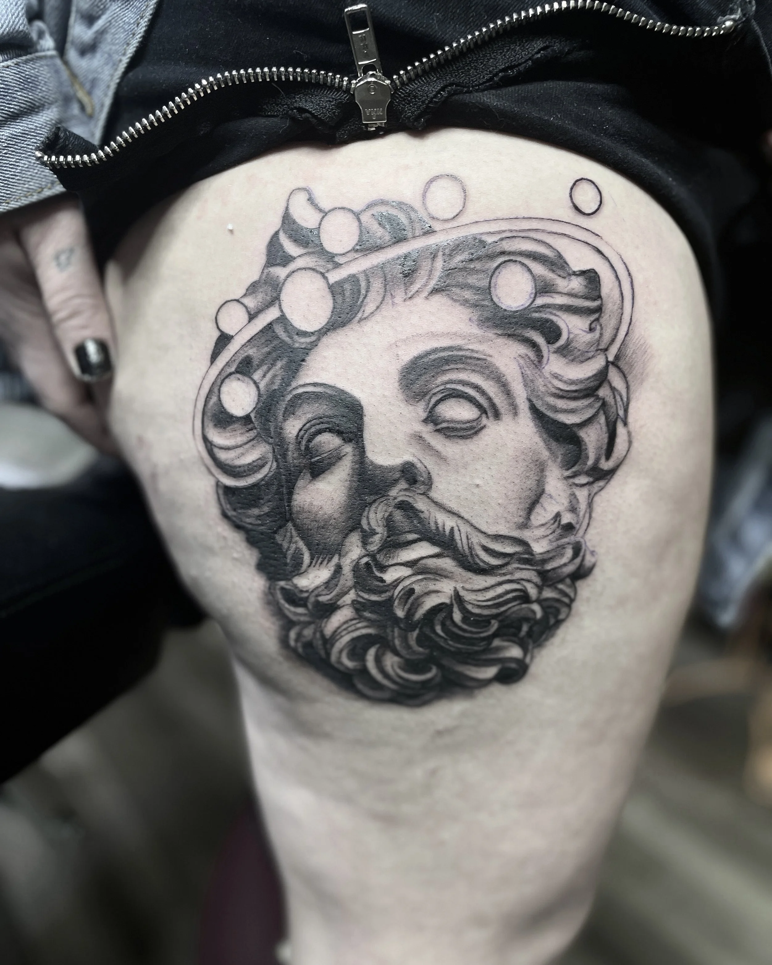 A black and gray tattoo of a classical statue's face with a beard and flowing hair, surrounded by abstract circular designs, on a person's thigh.