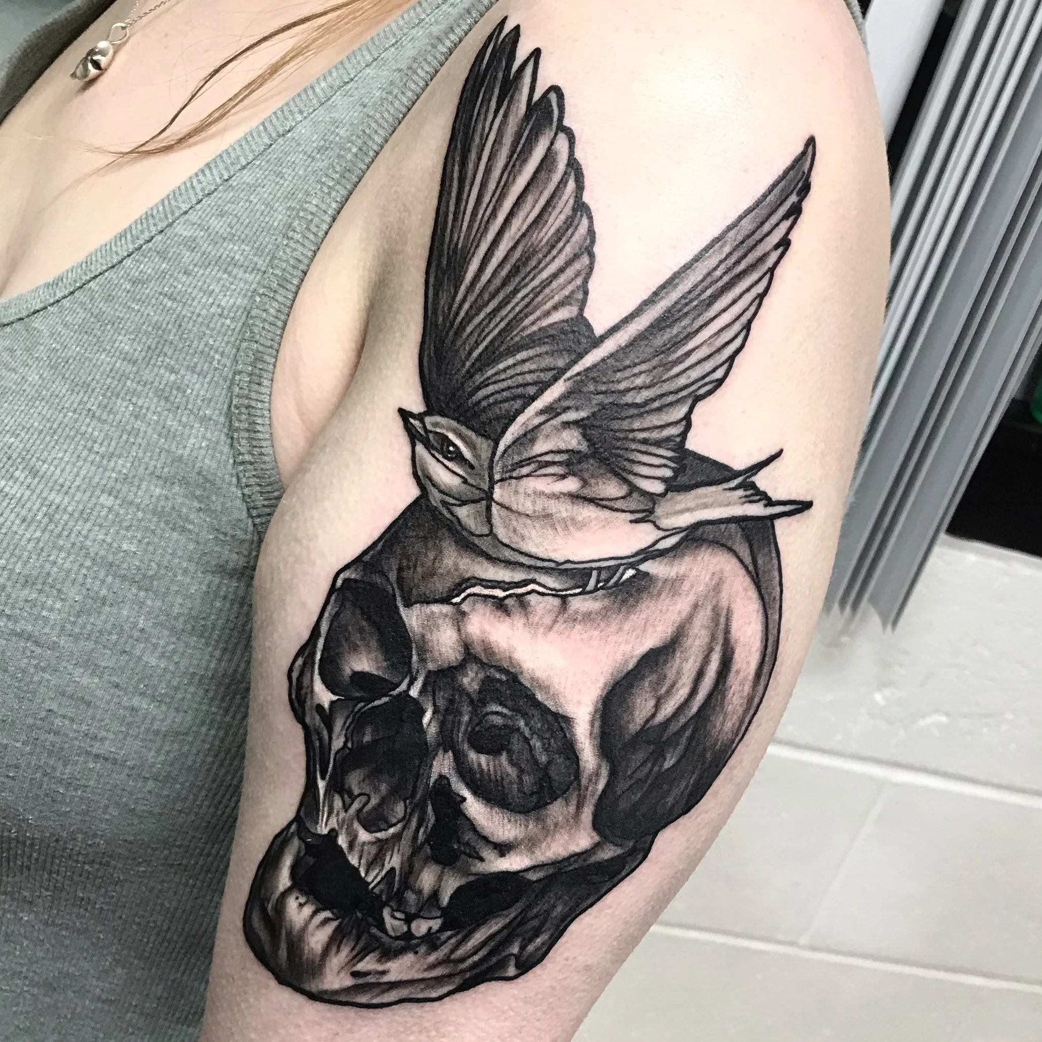 Black and gray tattoo of a skull with a bird flying above it on a person's upper arm.