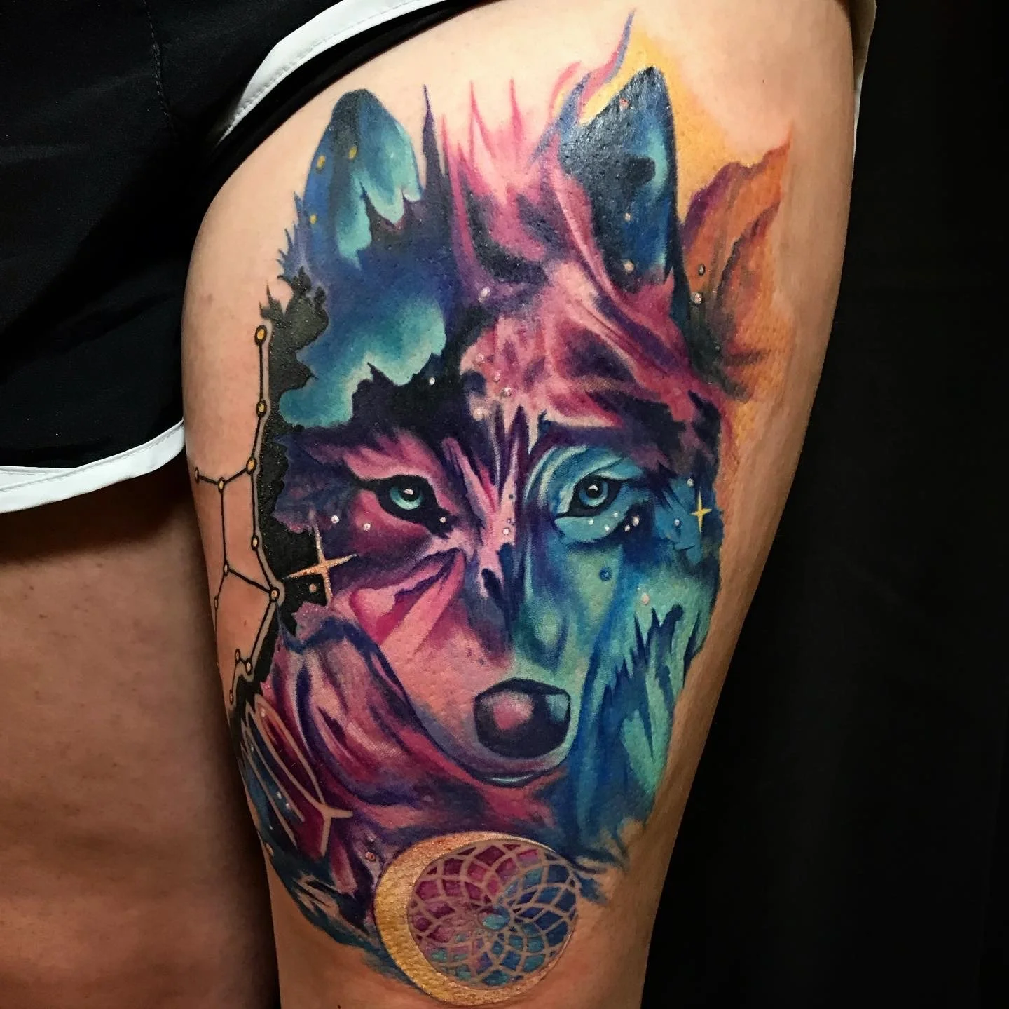 Colorful tattoo of a wolf's face on a person's upper arm, featuring celestial and cosmic elements with vibrant blues, pinks, and purples, including a moon and star designs.