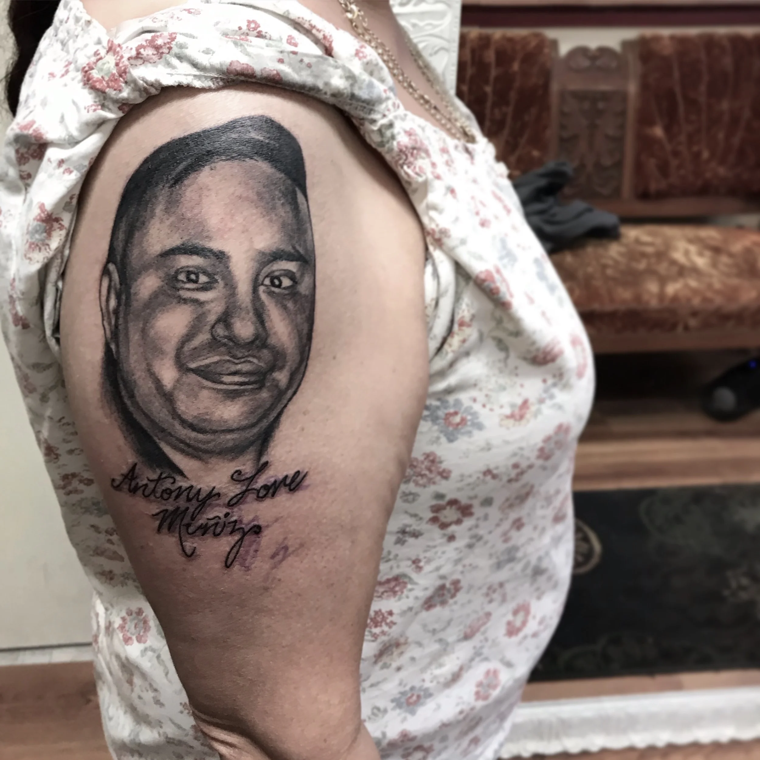 Tattoo of a realistic portrait of a smiling man with short hair on a person's upper arm, with the words "Anthony Love Mierz" written below in cursive.