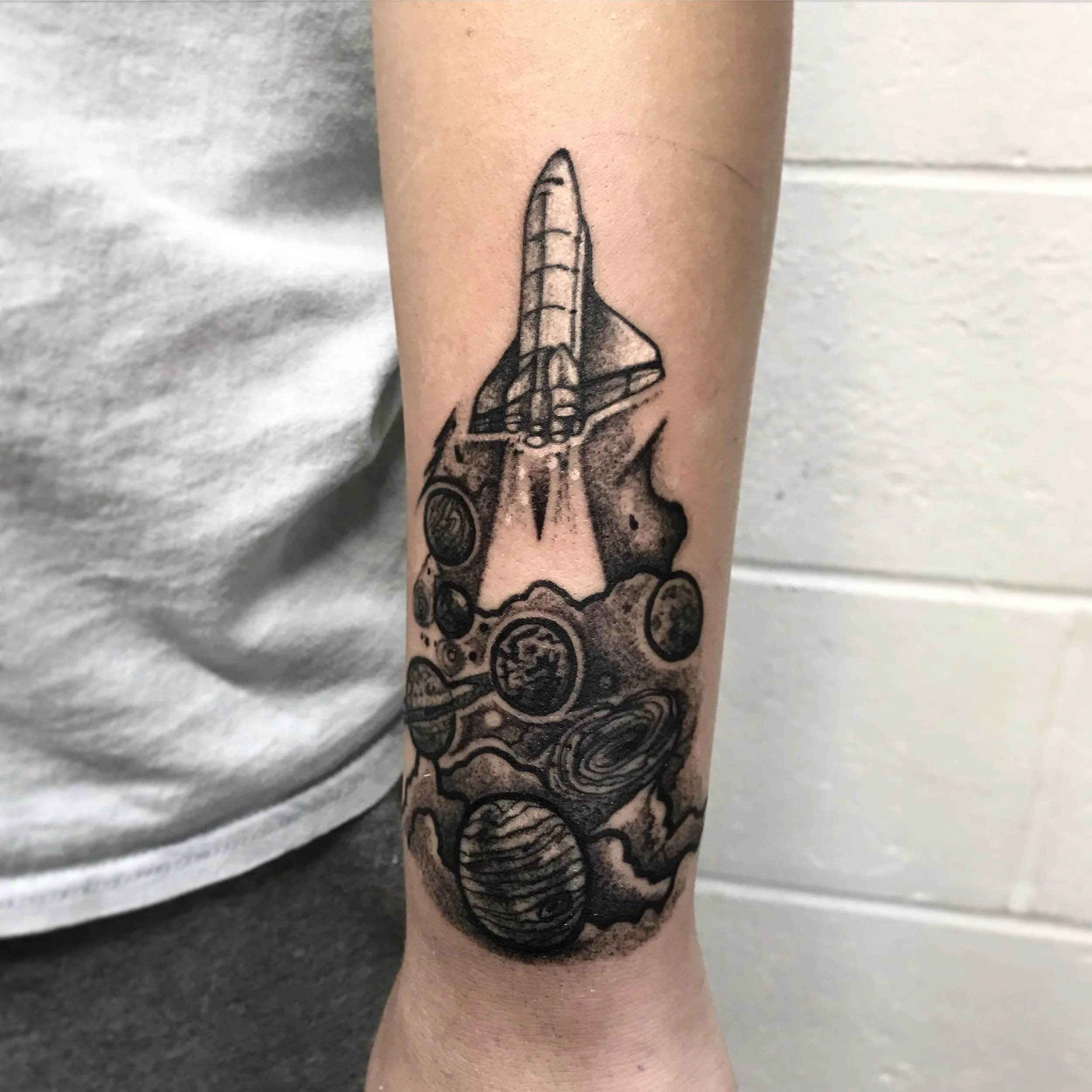 Tattoo of a space shuttle launching, with planets, stars, and abstract cosmic elements in black ink on a person's forearm.