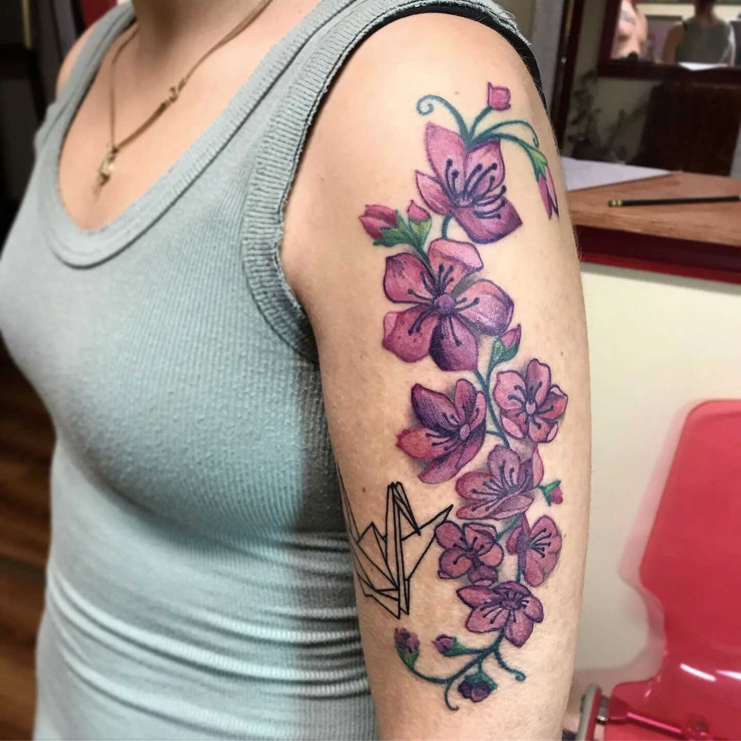 A person with a colorful tattoo of pink and purple flowers with green leaves and stems on their upper arm, next to a geometric origami-style design.