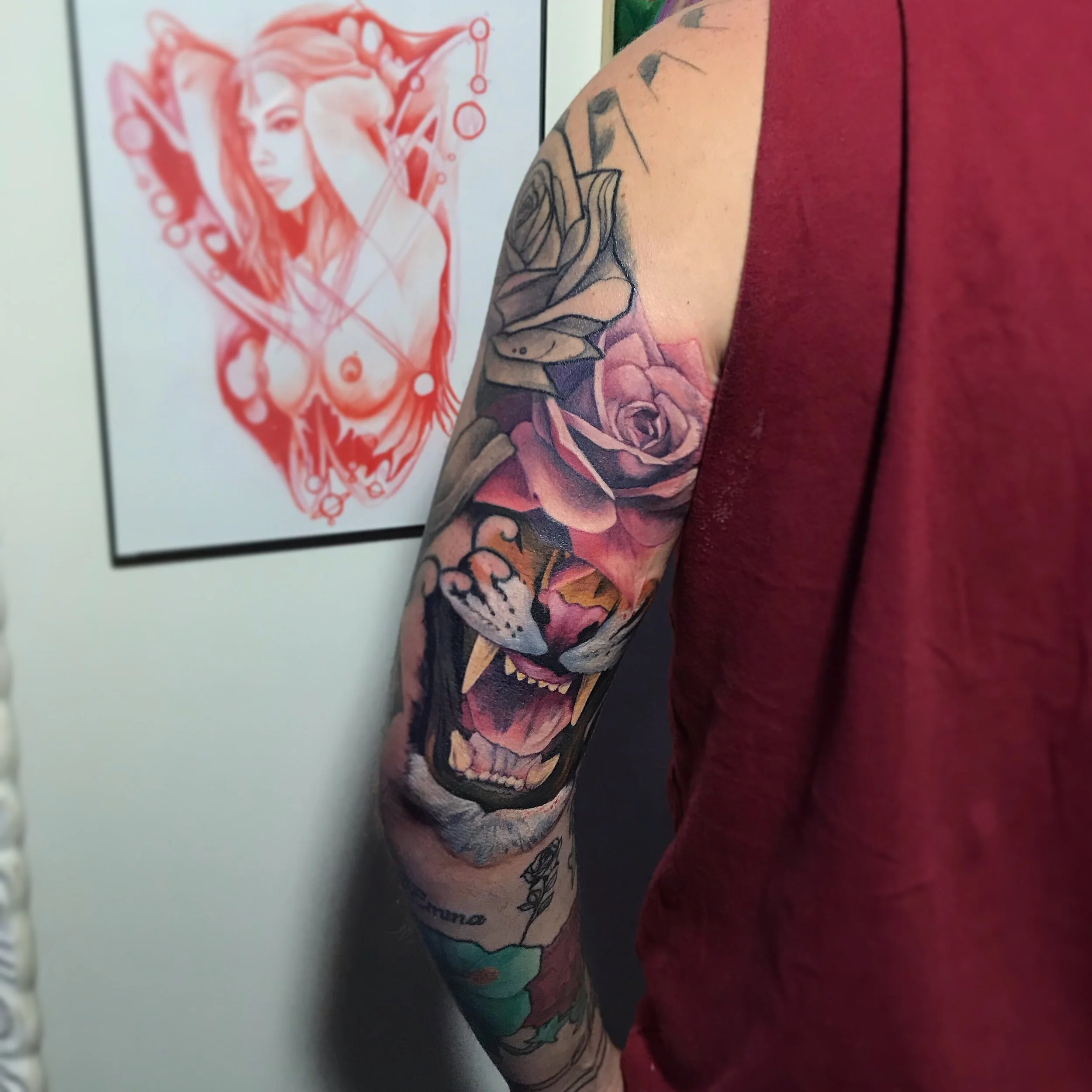Tattoo sleeve on a person's arm featuring a large snarling animal face, a purple rose, and other floral elements, with a framed artwork of a female elf doll hanging on the wall in the background.