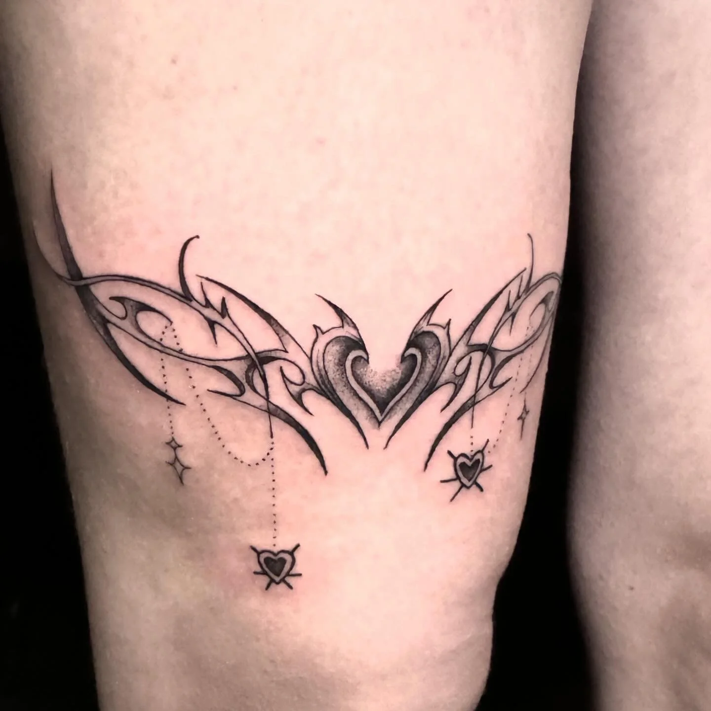 fine line  tattoo of a stylized heart with wing-like designs on the chest, with smaller heart and star motifs hanging from it.