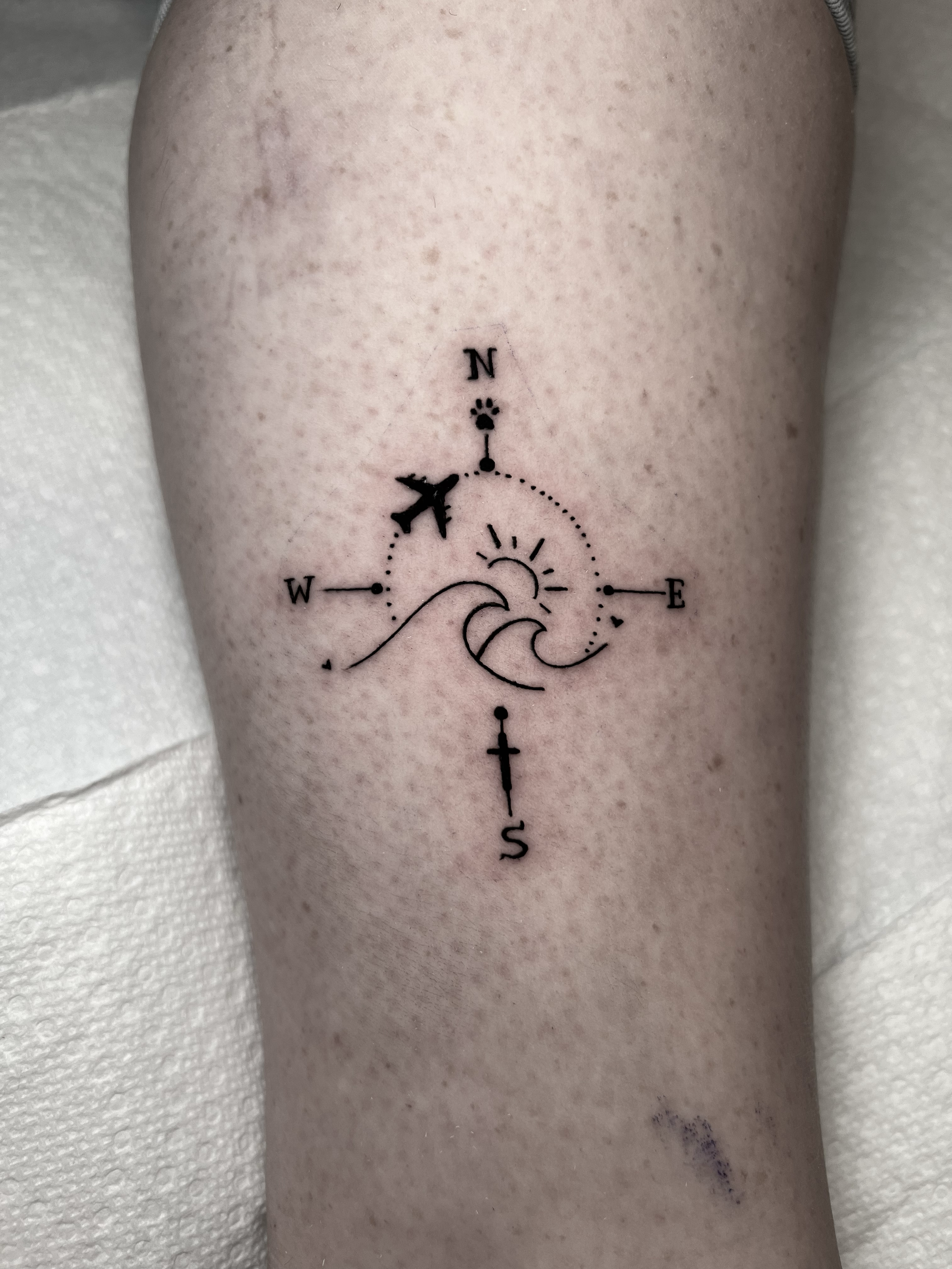 Tattoo of a fine line compass with cardinal directions, a moon and sun symbol, an airplane, and a sword on a person's thigh.