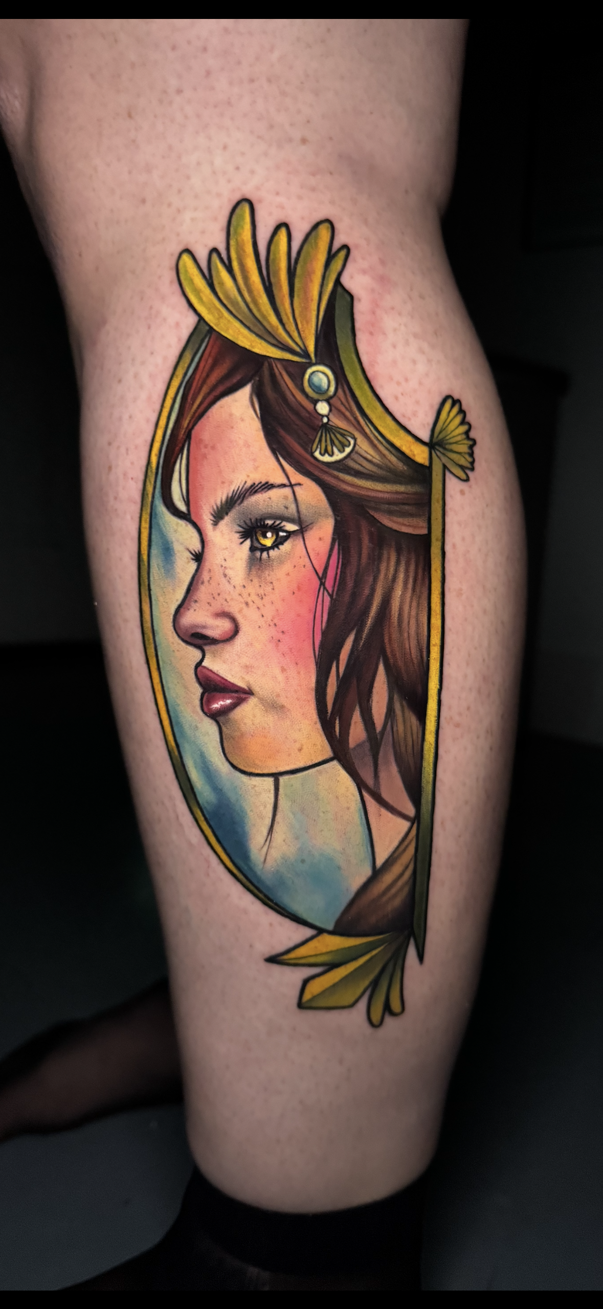 Colorful tattoo of a woman's profile with brown hair, freckles, yellow eye, and a yellow and green feathered headdress, set within an ornate frame.