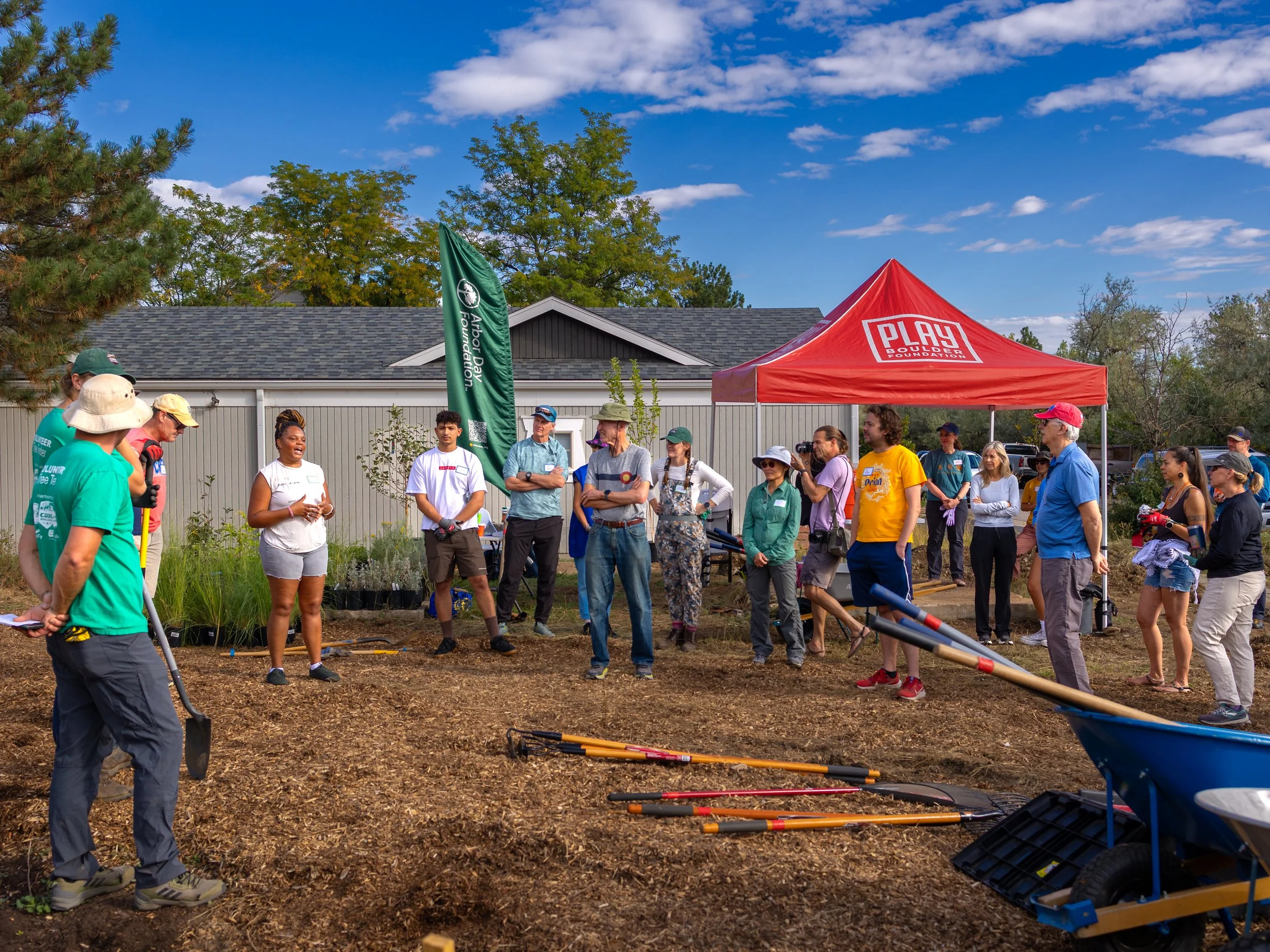 Group of people standing and listening to a speaker outdoors, shovels and wheelbarrows nearby. Clear day, partly cloudy.