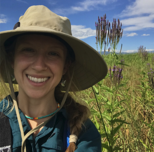 A young woman wearing a broad-rimmed sun hat with a natural landsape behind her.
