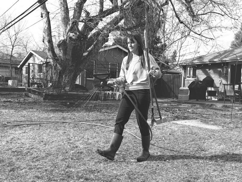 A black & white photo of a woman holding lawn tools in one hand and watering with the other hand. 