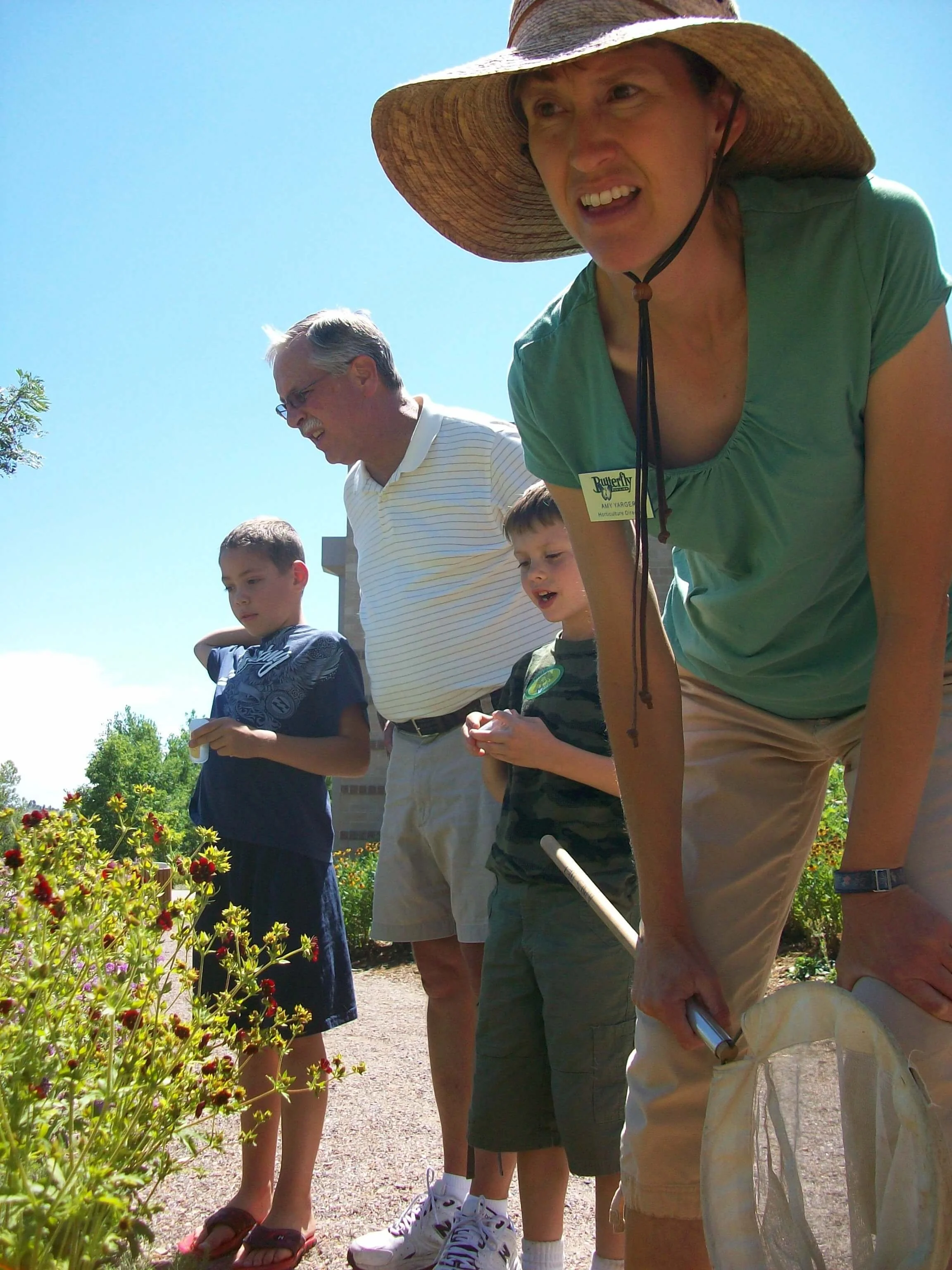 Amy Yarger in a straw hat teaching a class outdoors with an elderly man and two boys, looking at flowers.