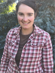 A young woman with long, wavy brown hair sitting outdoors on the ground covered with fallen autumn leaves, smiling at the camera.