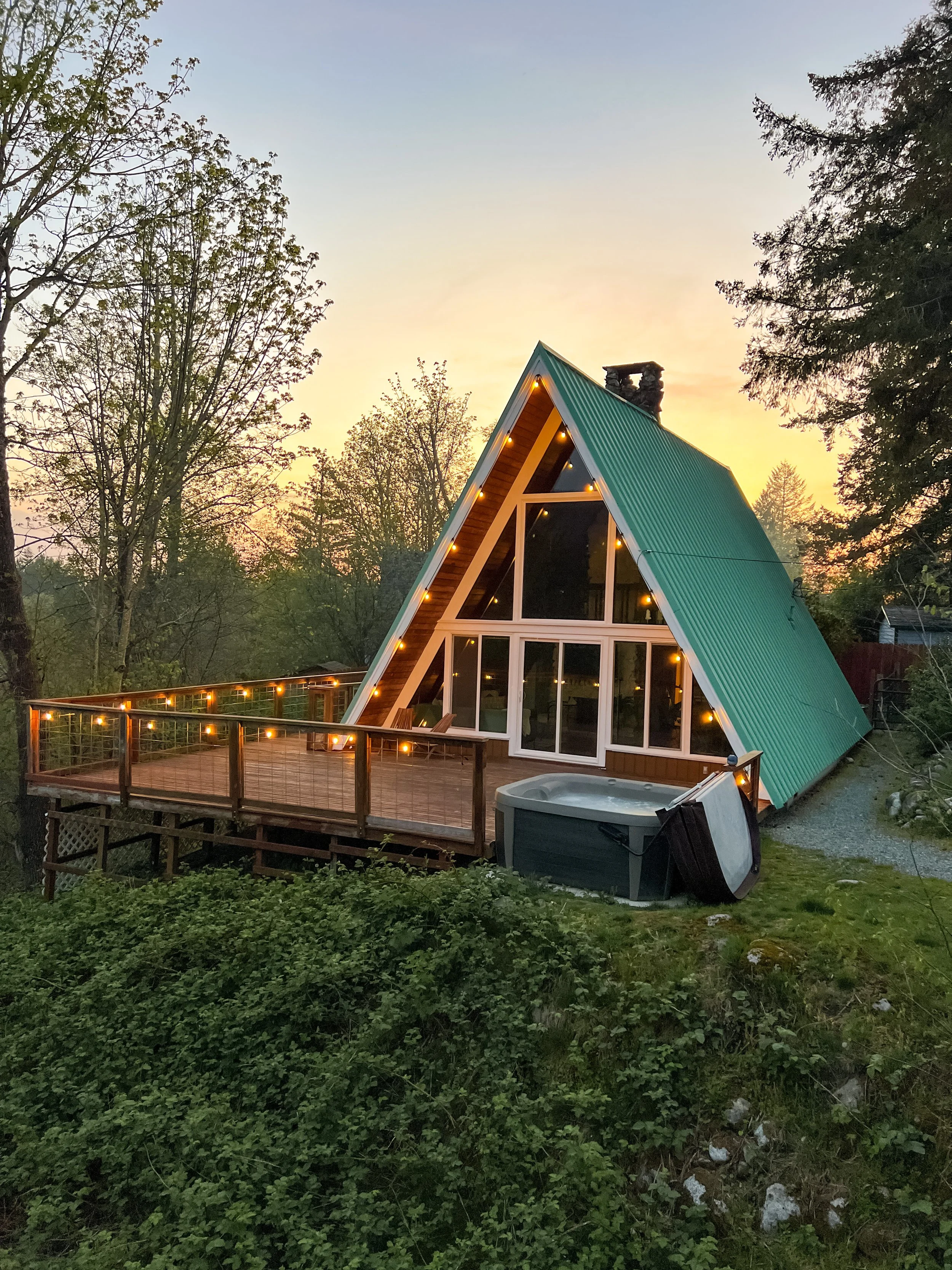 A-frame cabin with large glass windows and a green metal roof, decorated with string lights on the deck, surrounded by trees at sunset, with a hot tub on the lawn in front.