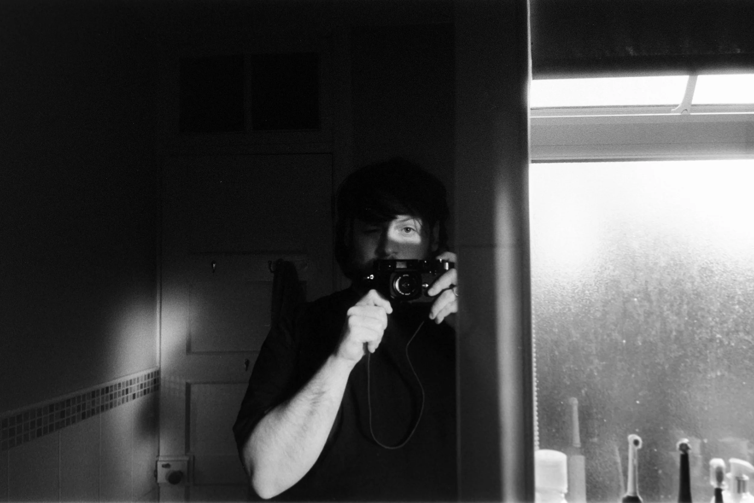 Black and white photo of a person taking a mirror selfie in a bathroom. The person is holding a camera up to their face, with uneven lighting illuminating one eye and part of their face. The bathroom has a frosted window, toothbrushes, and bathroom fixtures visible.